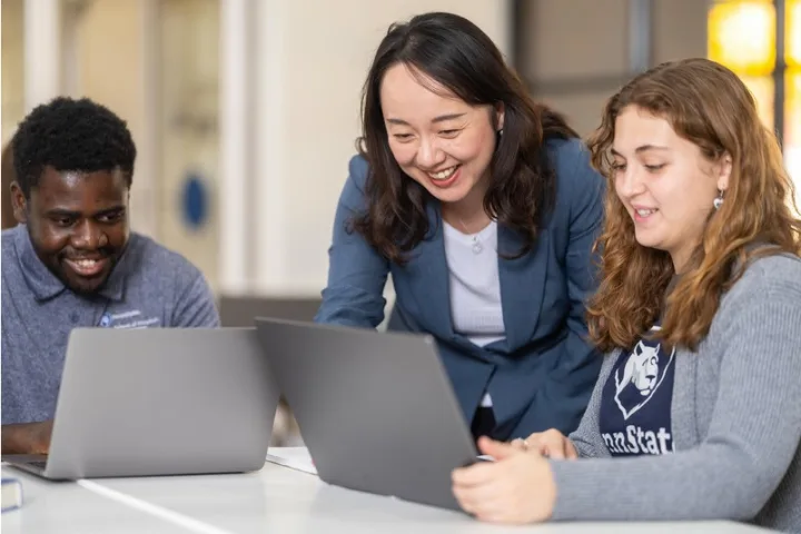 Professor helps with two students at a table with laptops