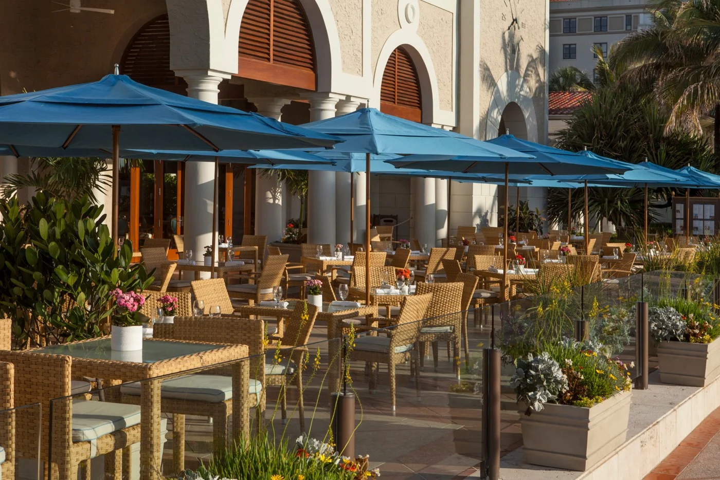 Outdoor patio dining area with blue umbrellas, wicker furniture, and Spanish-style architecture.