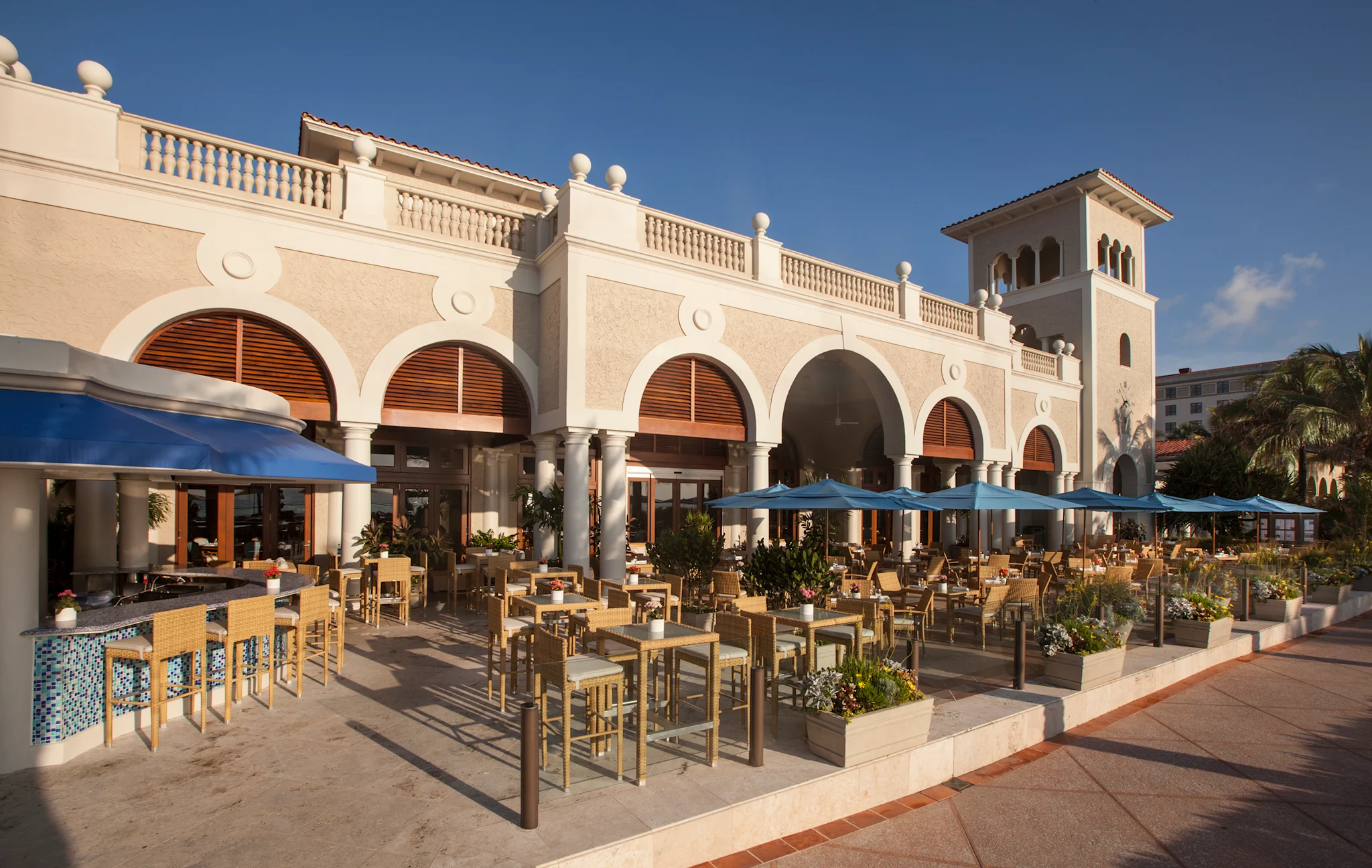 Mediterranean-style resort with beige arched facade, outdoor patio dining with blue umbrellas and wooden furniture.