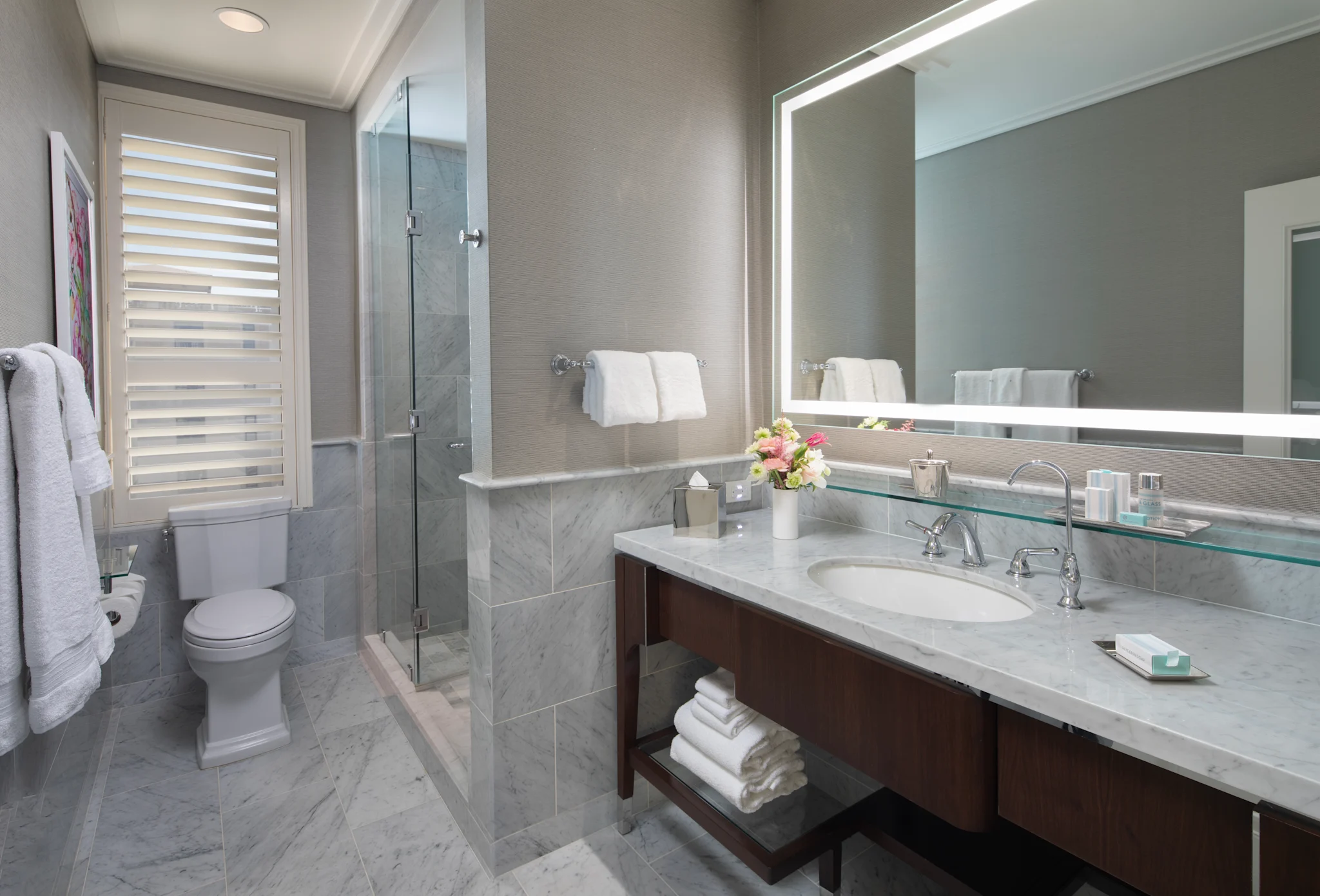 Modern bathroom with gray marble vanity, backlit mirror, glass shower, and white towels.