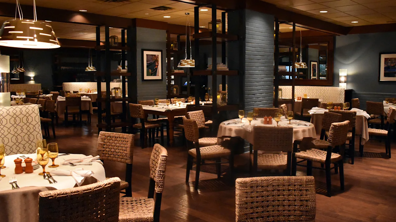 Upscale restaurant interior with wicker chairs, white tablecloths, pendant lighting, and dark walls.