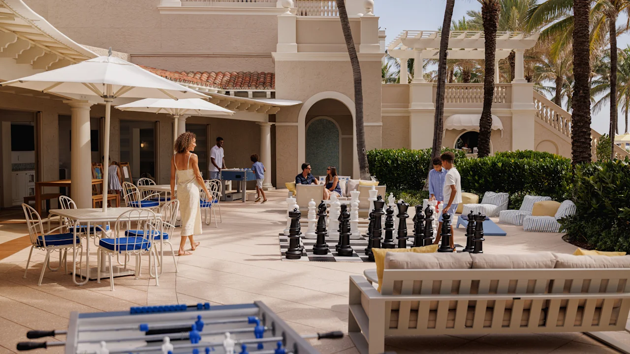 Luxury resort courtyard with palm trees, outdoor chess set, and guests relaxing on patio furniture.