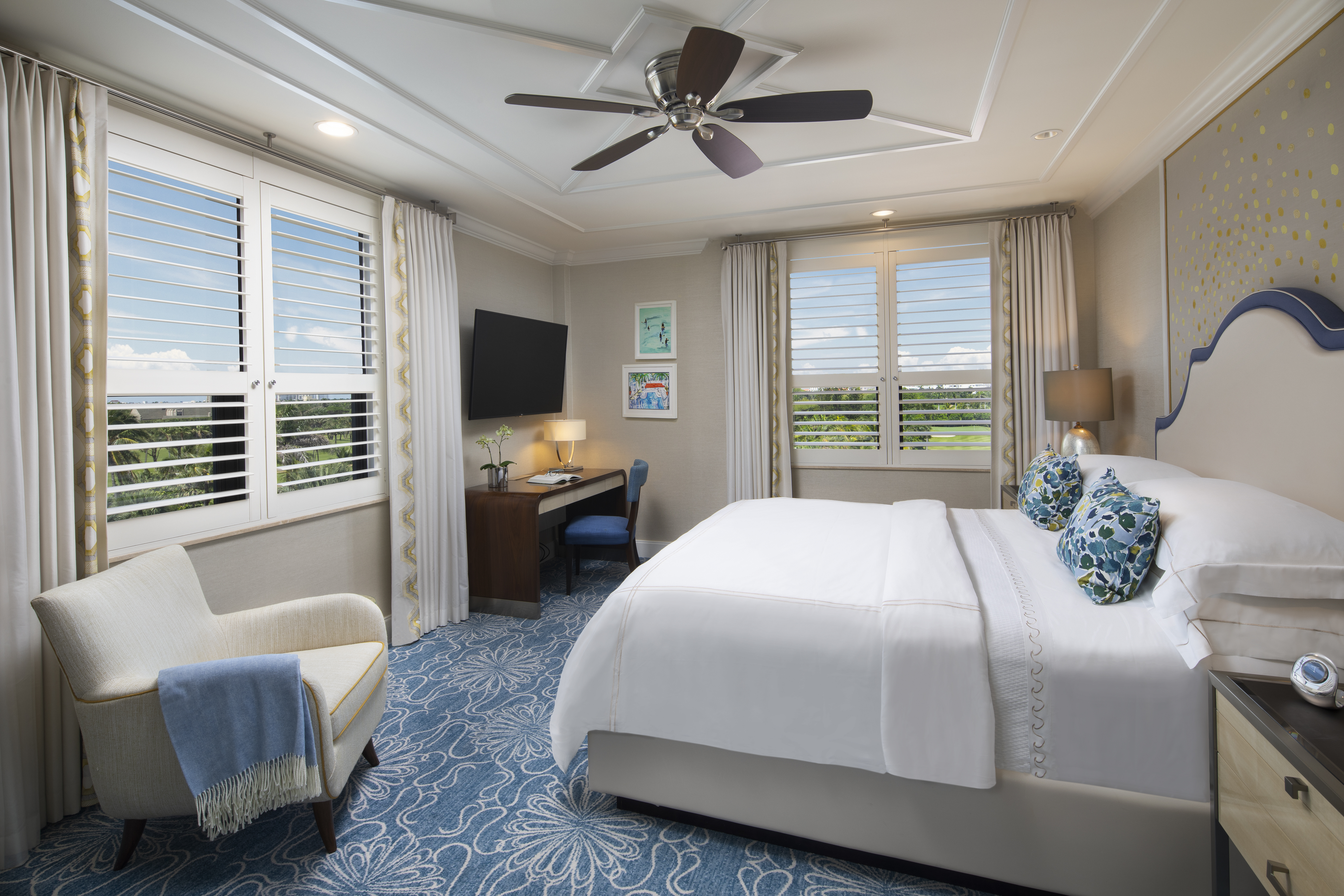 Elegant hotel bedroom with white bedding, blue patterned carpet, ceiling fan, and plantation shutters.