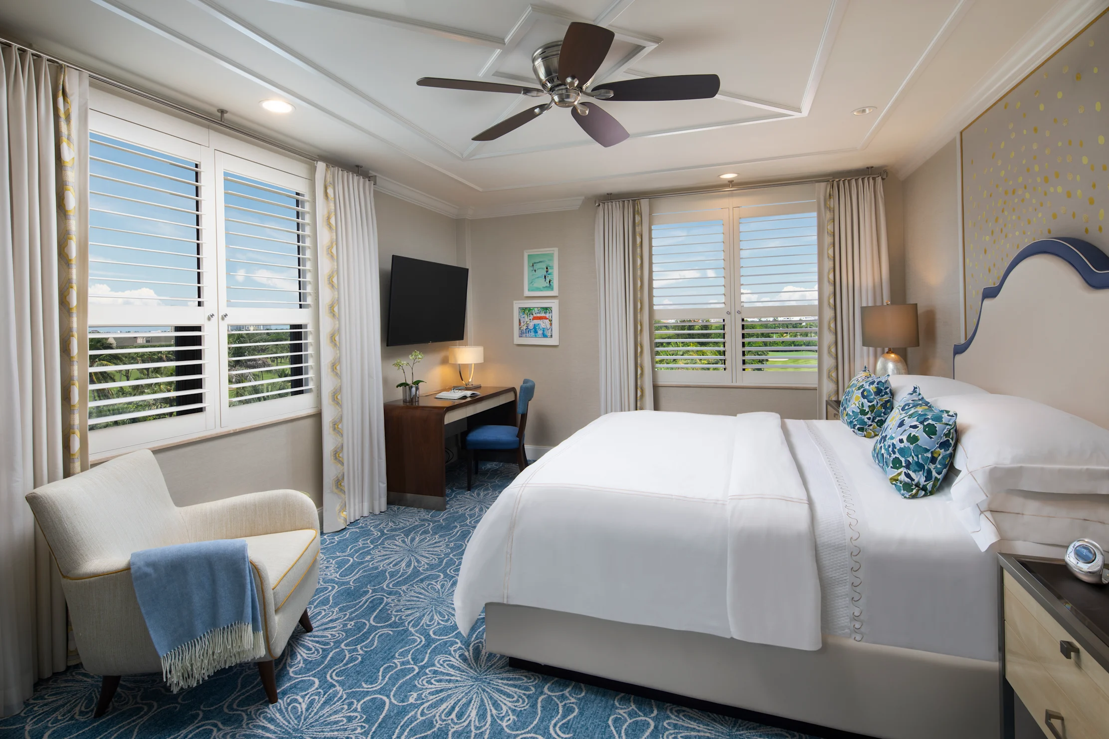 Elegant hotel bedroom with white bedding, blue patterned carpet, ceiling fan, and plantation shutters.