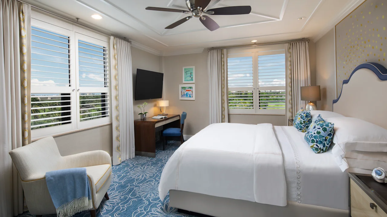 Elegant hotel bedroom with white bedding, blue patterned carpet, ceiling fan, and plantation shutters.
