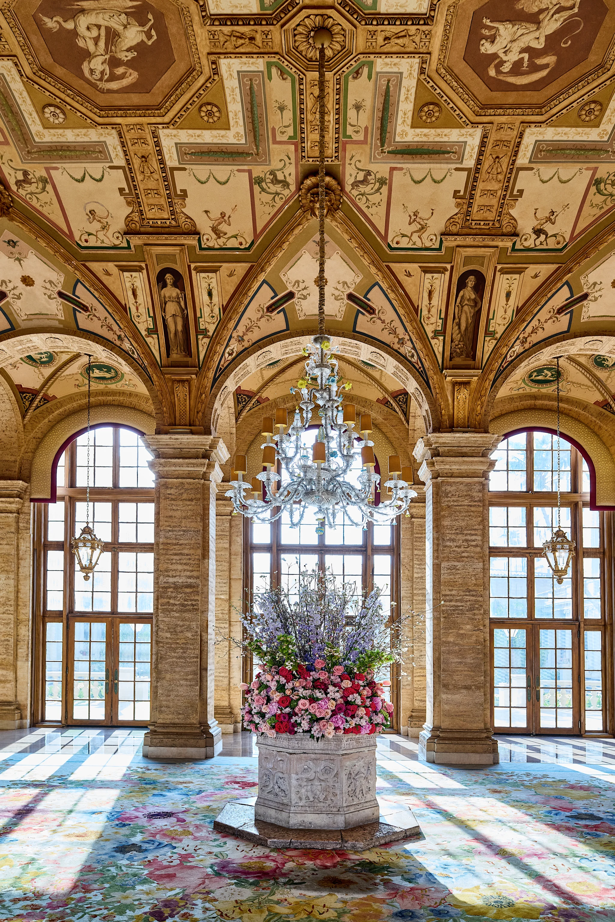 Ornate palace interior with frescoed vaulted ceiling, crystal chandelier, stone columns, and floral arrangement.