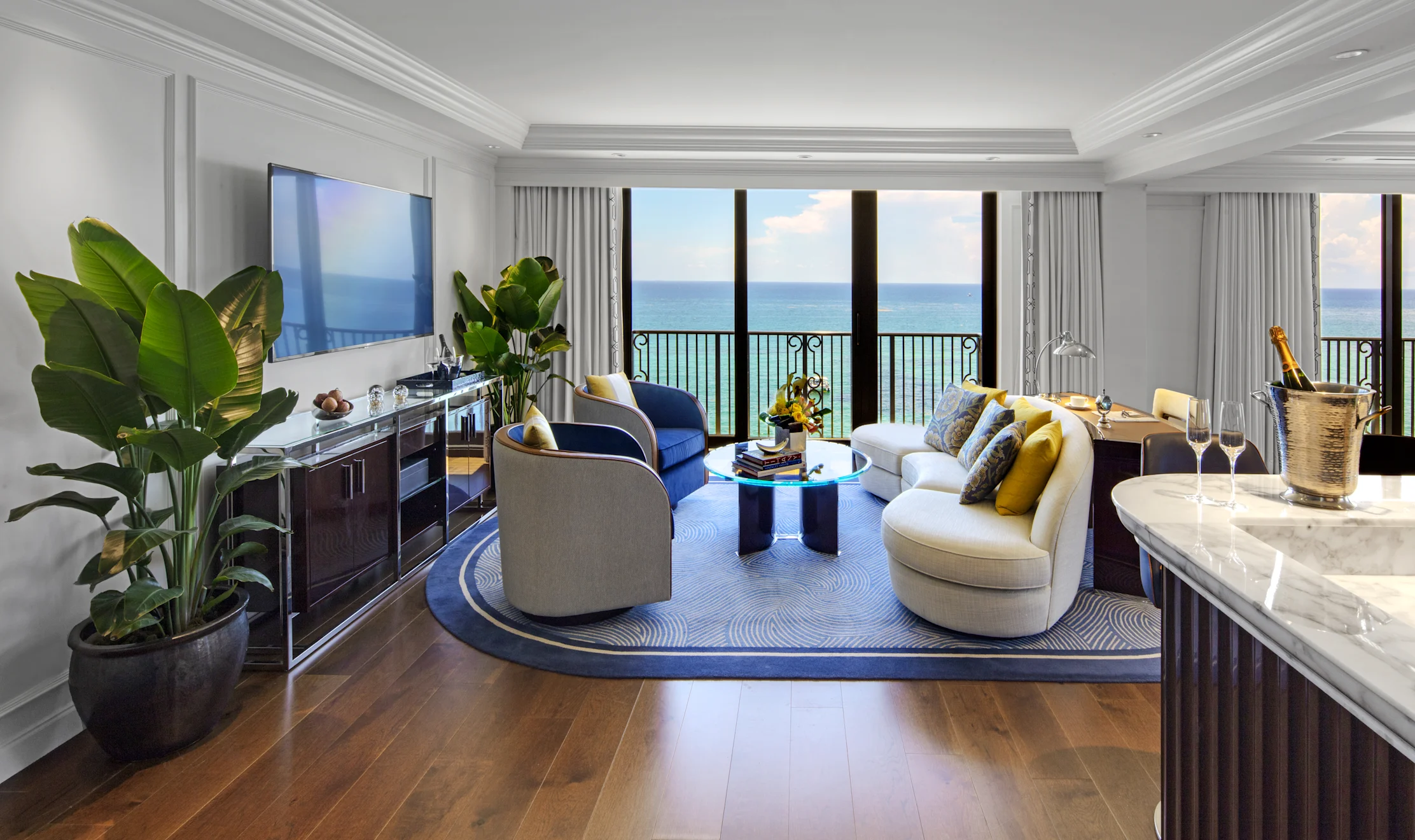 Luxury hotel suite with ocean view, modern furniture, blue accents, and champagne on marble counter.