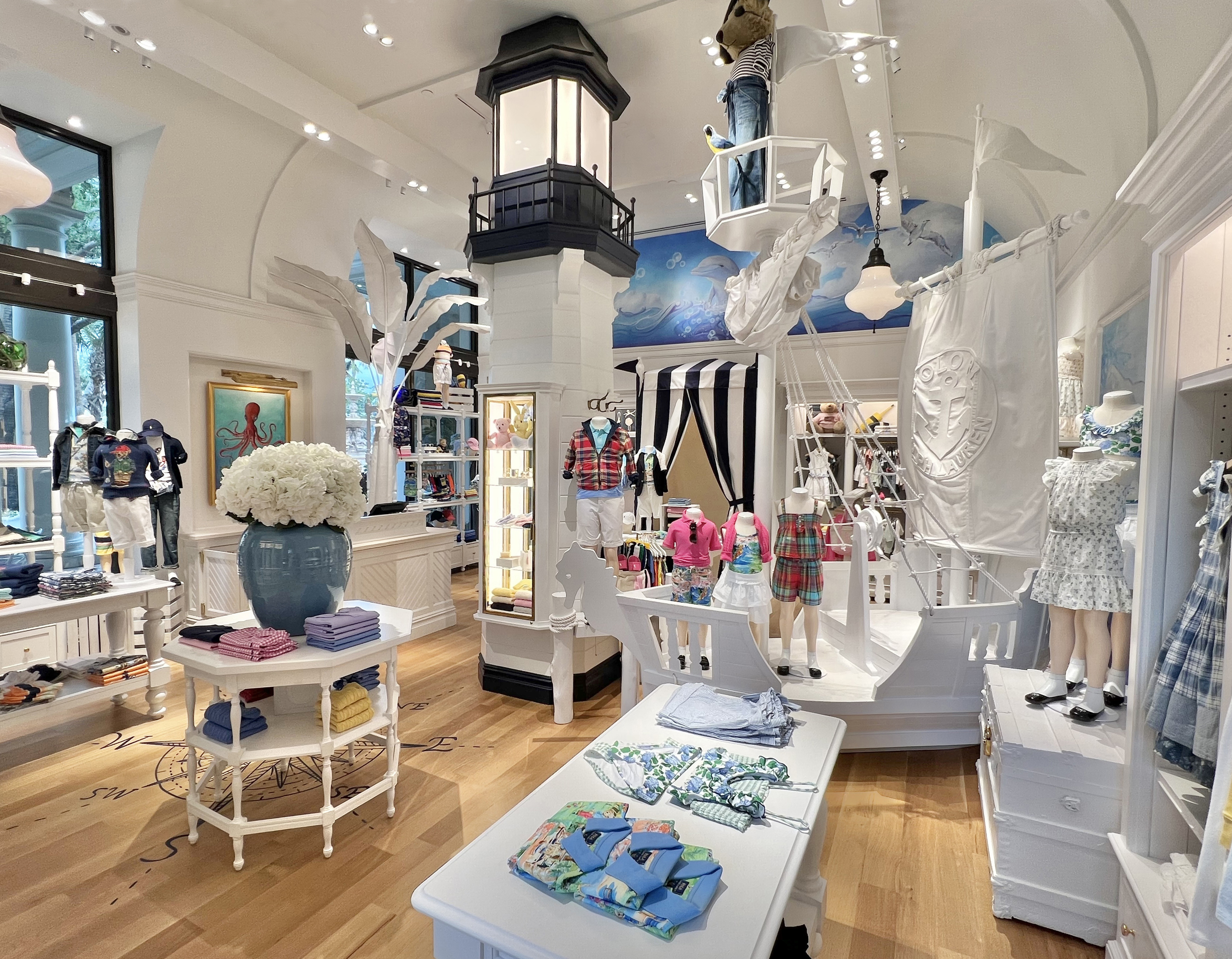 Nautical-themed children's clothing store with white decor, lighthouse column, ship figurehead, and colorful apparel displays.