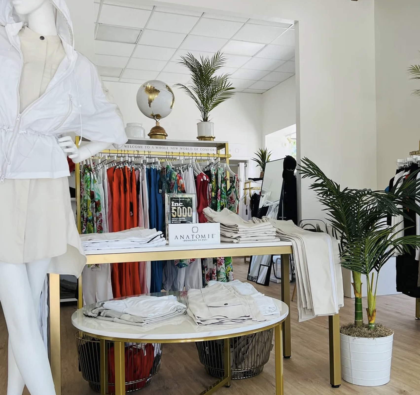 Bright boutique interior with colorful clothing racks, white display table, globe decor, and plants.