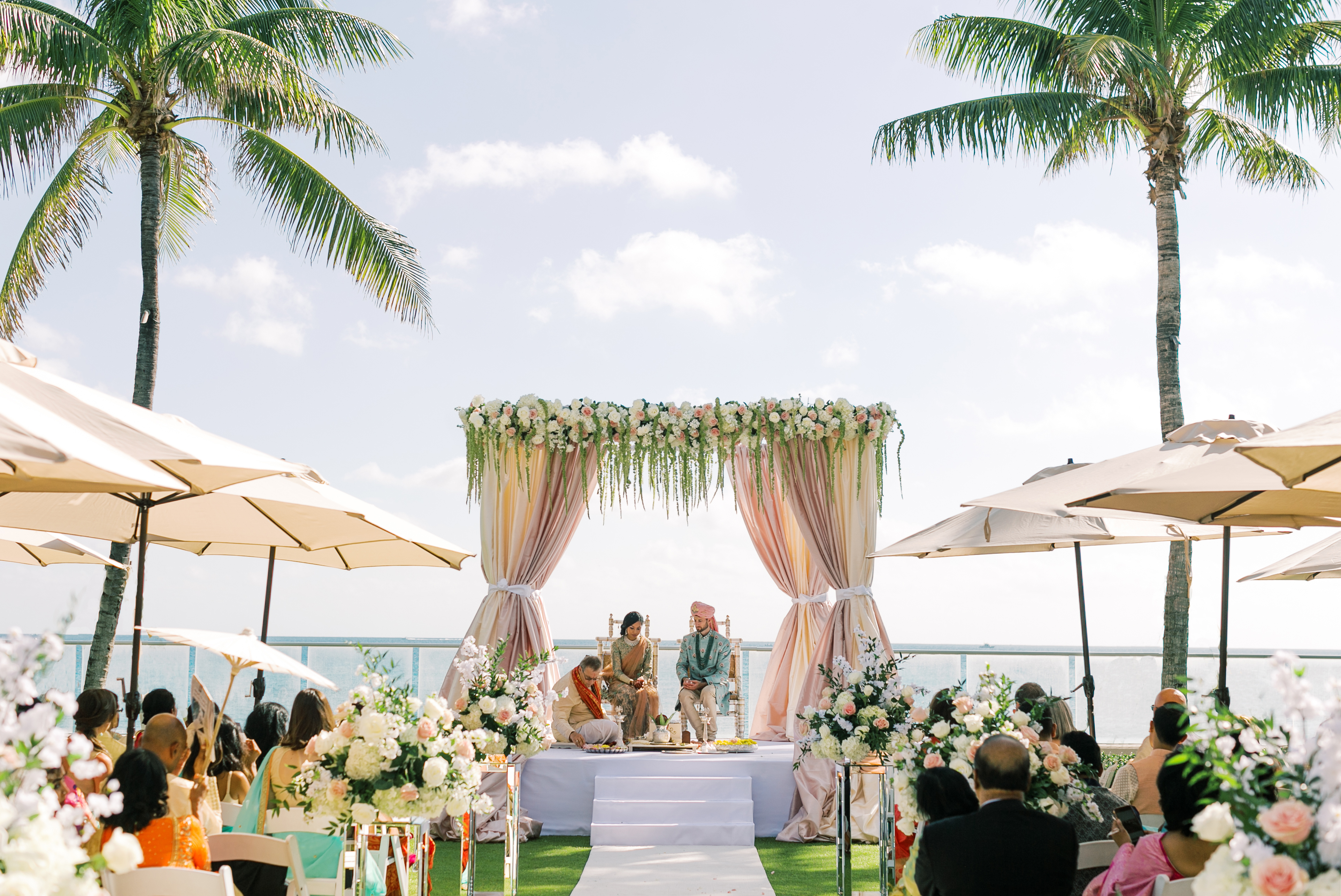 Outdoor beach wedding ceremony with decorated floral arch, palm trees, and guests under umbrellas.