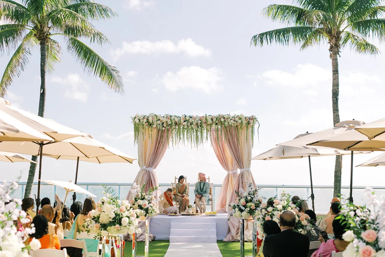 Outdoor beach wedding ceremony with decorated floral arch, palm trees, and guests under umbrellas.