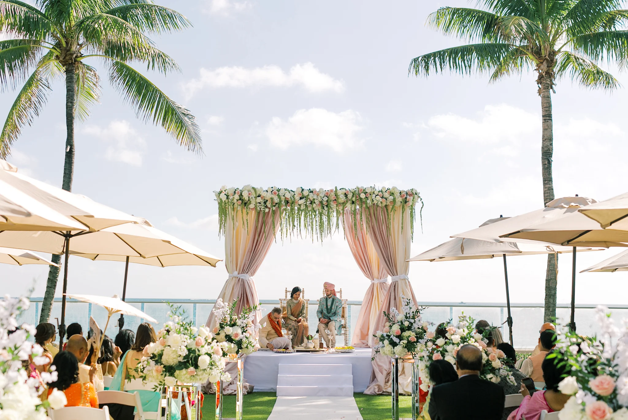 Outdoor beach wedding ceremony with decorated floral arch, palm trees, and guests under umbrellas.