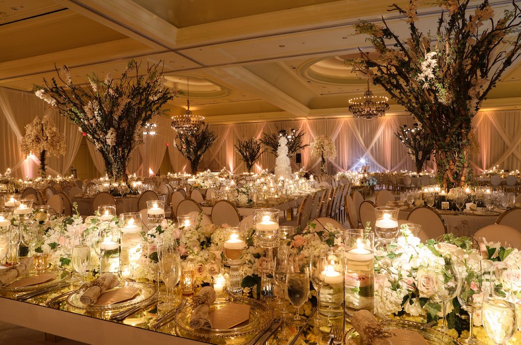 Elegant wedding reception ballroom with tall floral centerpieces, candles, draped fabric, and chandeliers.