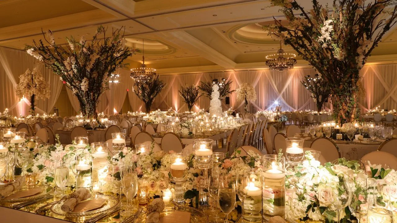 Elegant wedding reception ballroom with tall floral centerpieces, candles, draped fabric, and chandeliers.