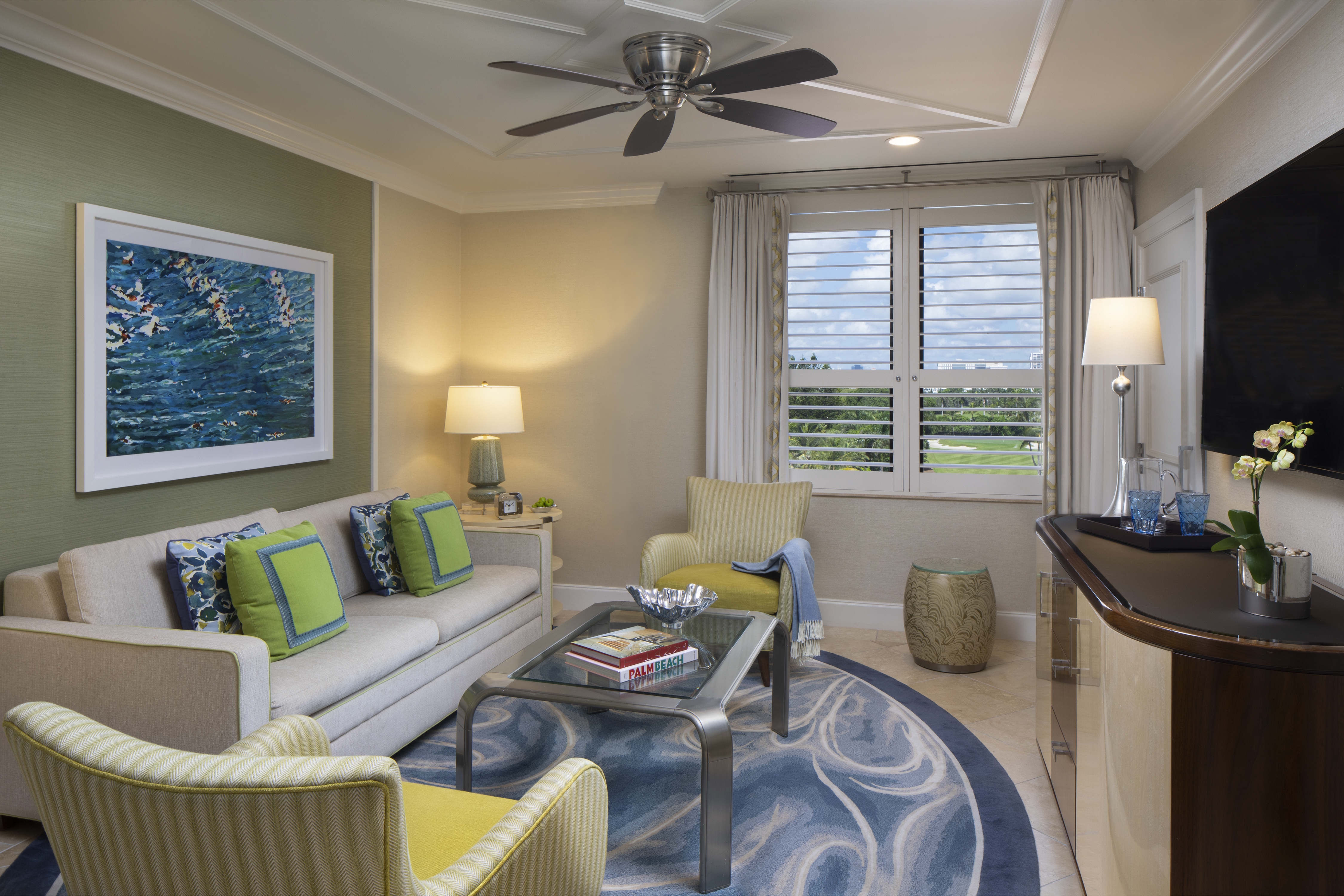 Modern living room with beige sofa, green accent chairs, ocean artwork, ceiling fan, and white shutters.