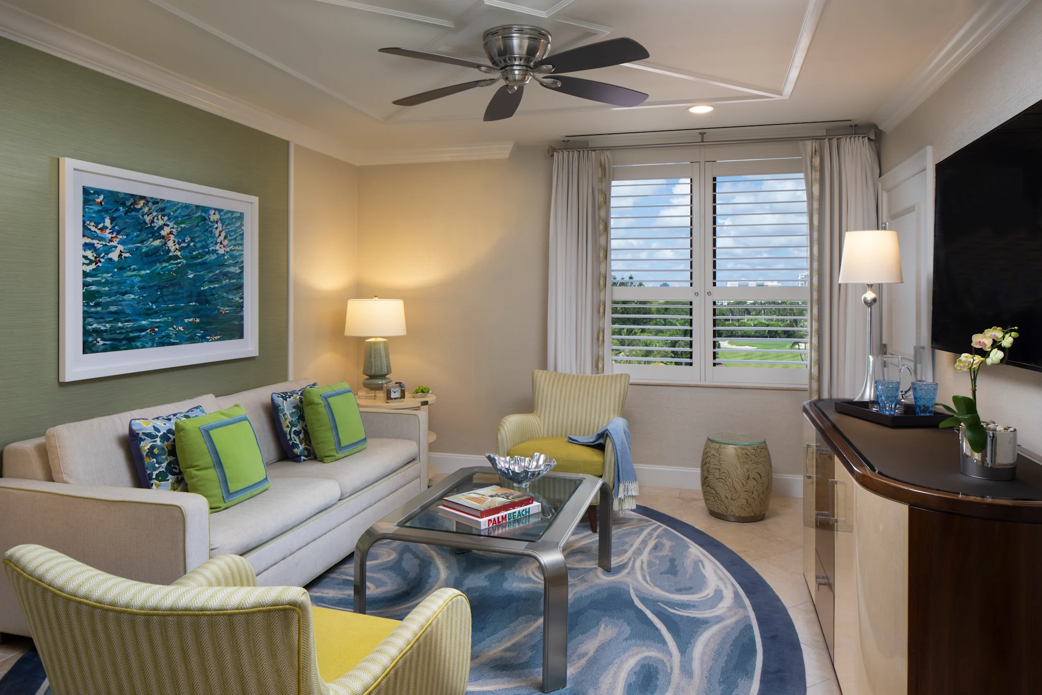 Modern living room with beige sofa, green accent chairs, ocean artwork, ceiling fan, and white shutters.