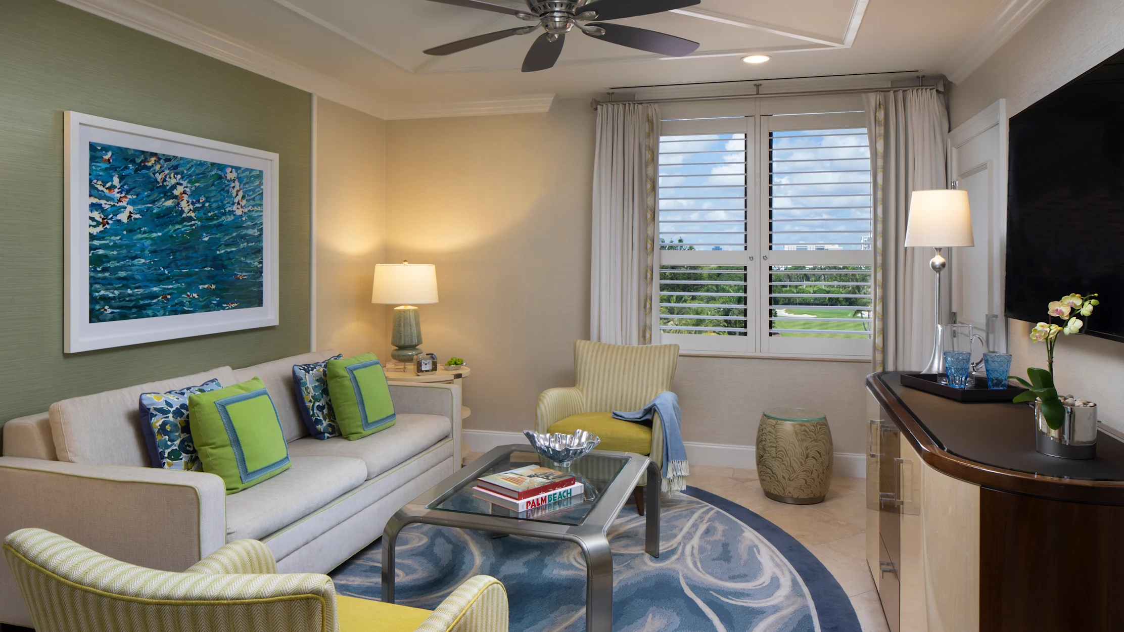 Modern living room with beige sofa, green accent chairs, ocean artwork, ceiling fan, and white shutters.