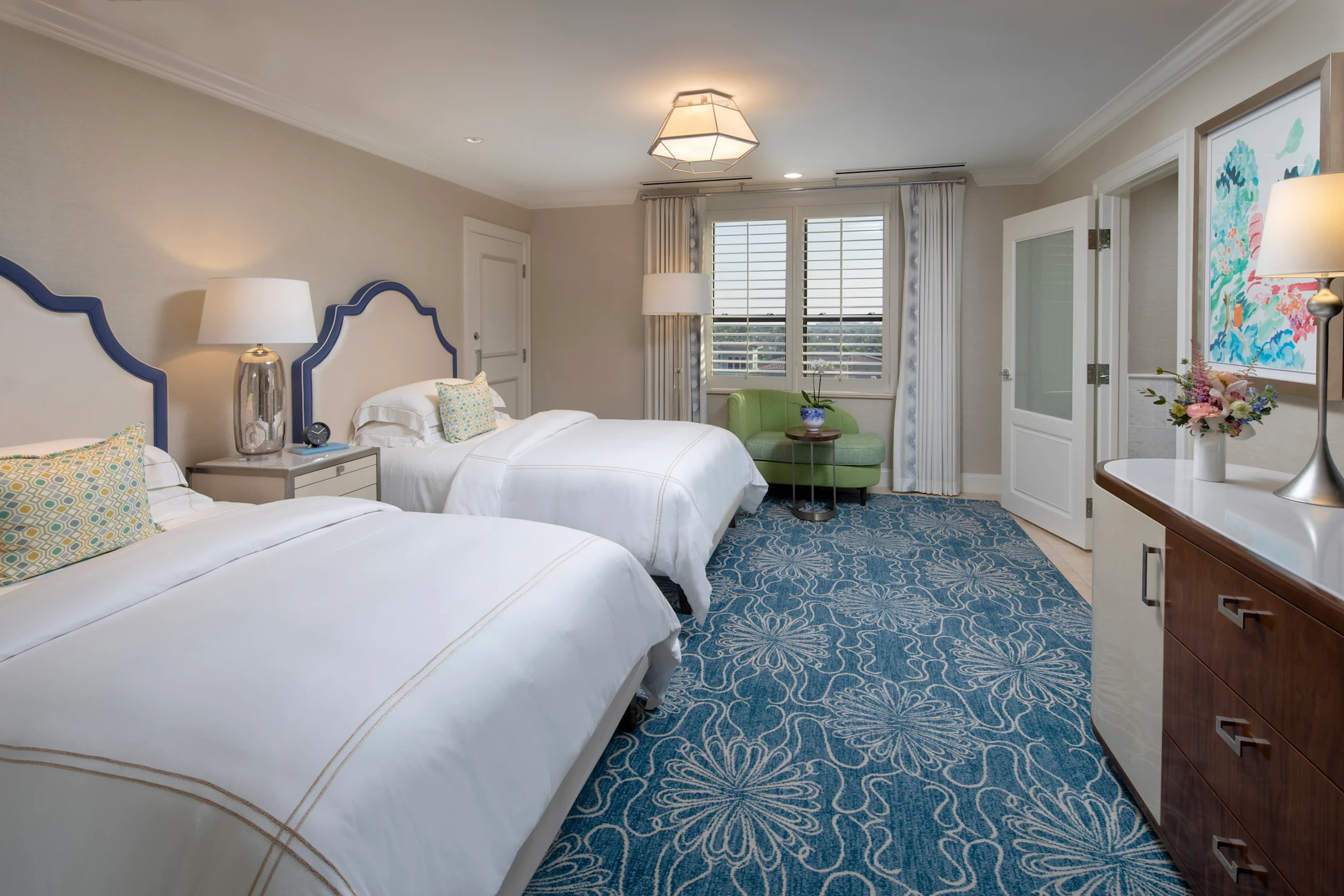 Elegant hotel bedroom with two white beds, blue patterned carpet, green accent chair, and coastal artwork.