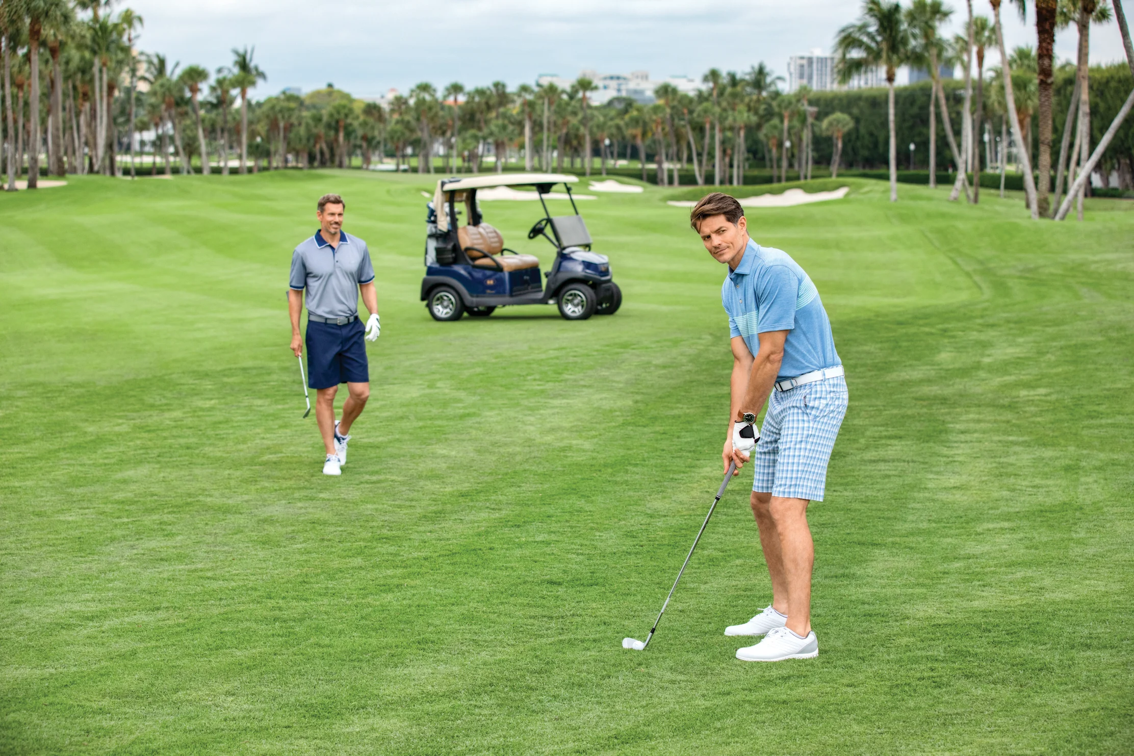 Two men playing golf on a lush green course with palm trees and a golf cart in the background.