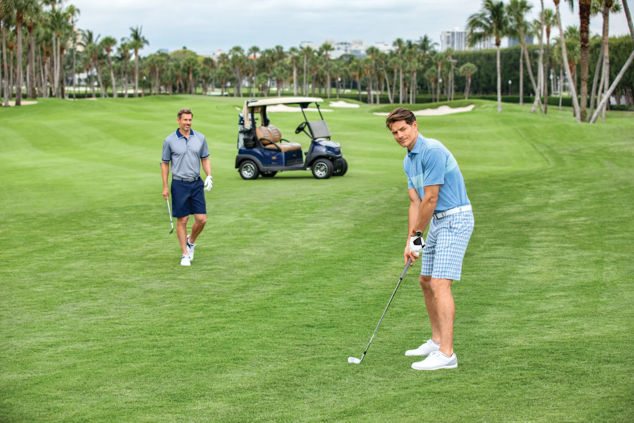 Two men playing golf on a lush green course with palm trees and a golf cart in the background.