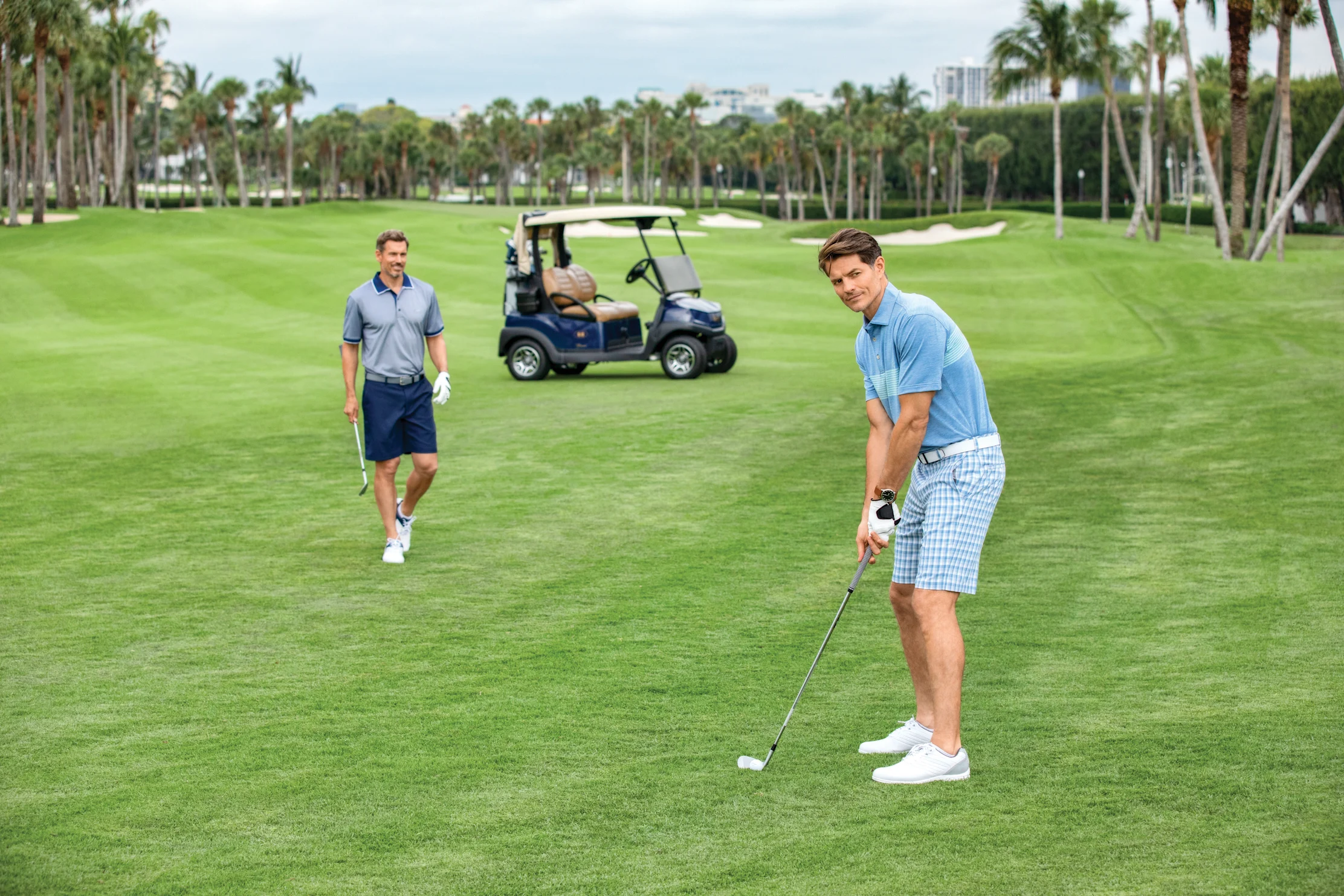 Two men playing golf on a lush green course with palm trees and a golf cart in the background.