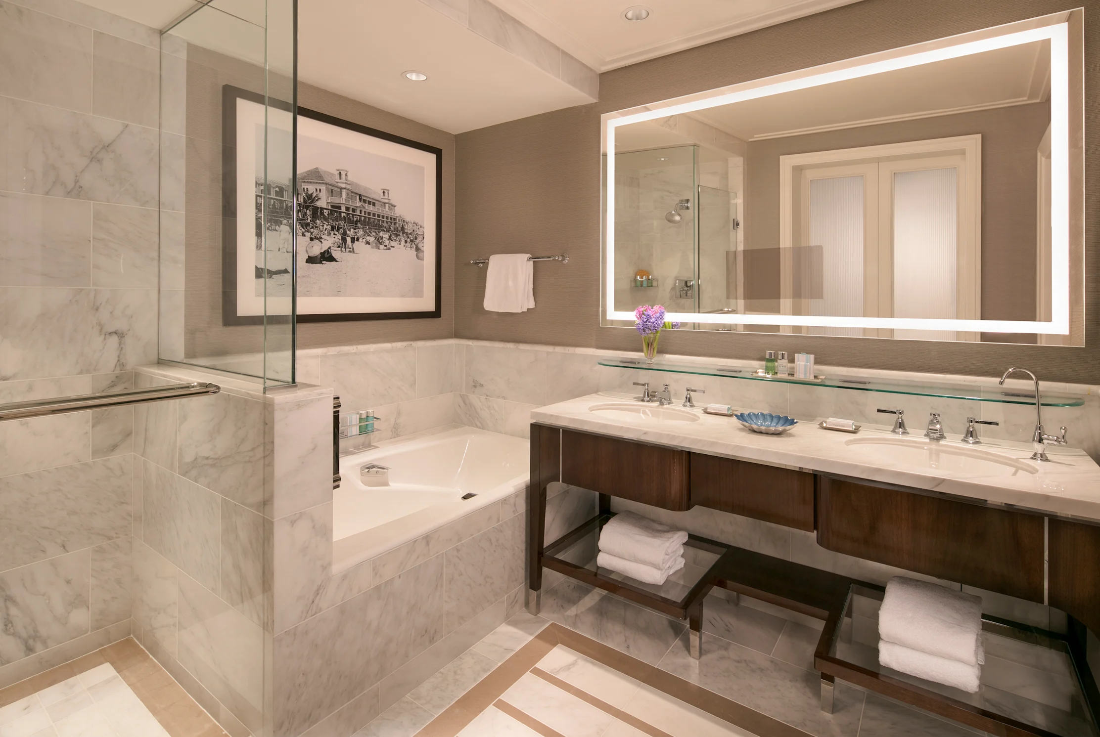 Luxury hotel bathroom with marble tub, double vanity, backlit mirror, and glass shower enclosure.