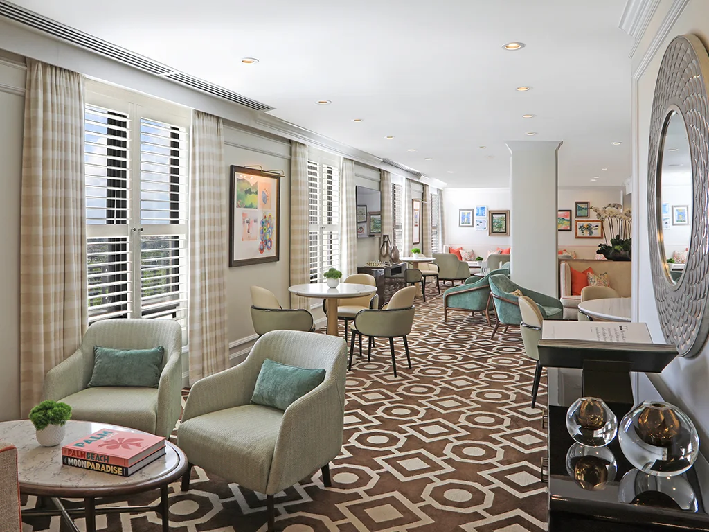 Elegant hotel lounge with beige armchairs, geometric patterned carpet, white shutters, and teal accents.