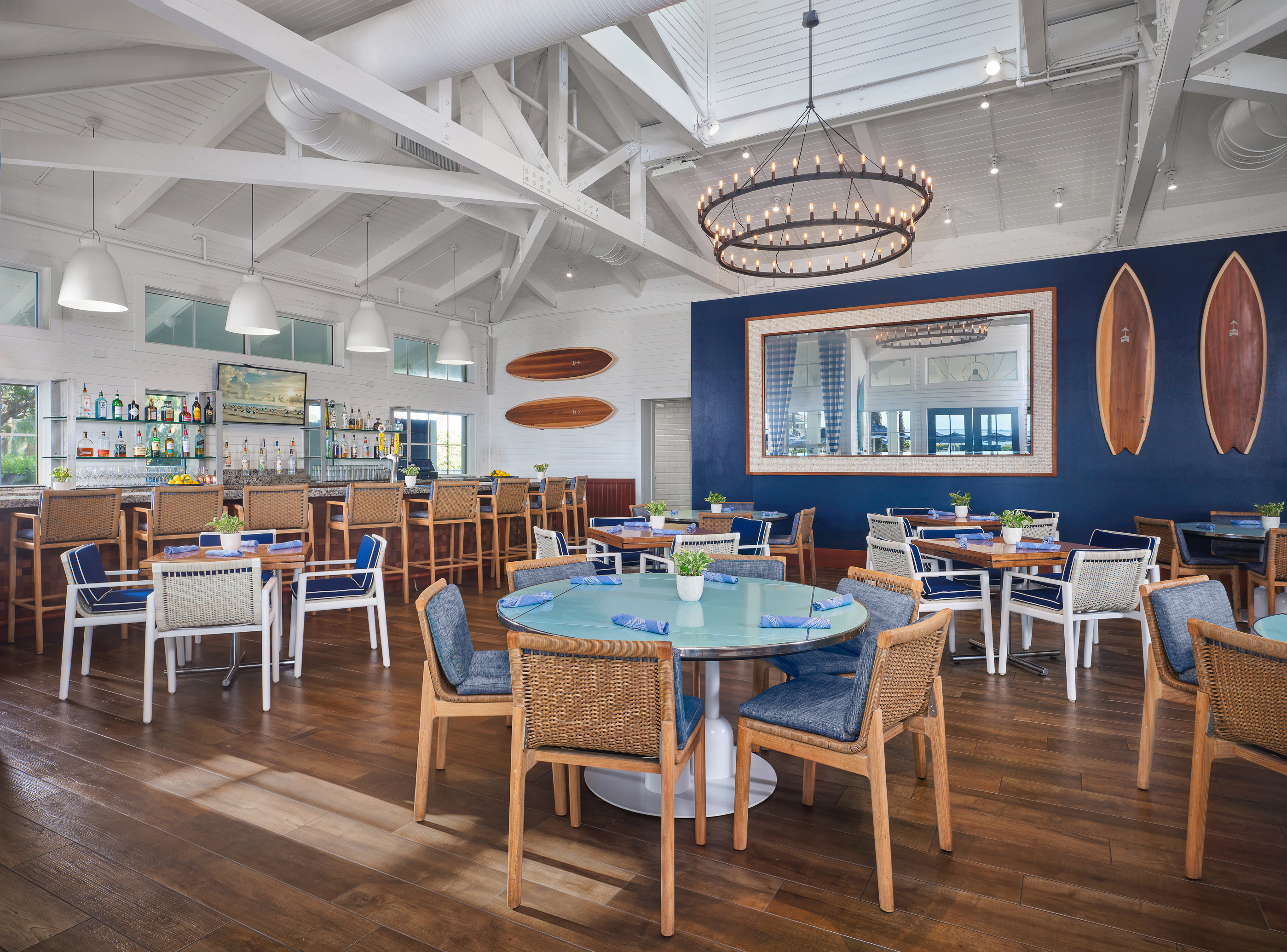 Modern coastal restaurant interior with vaulted ceilings, circular chandelier, bar, coral booth seating, and surfboard decor.