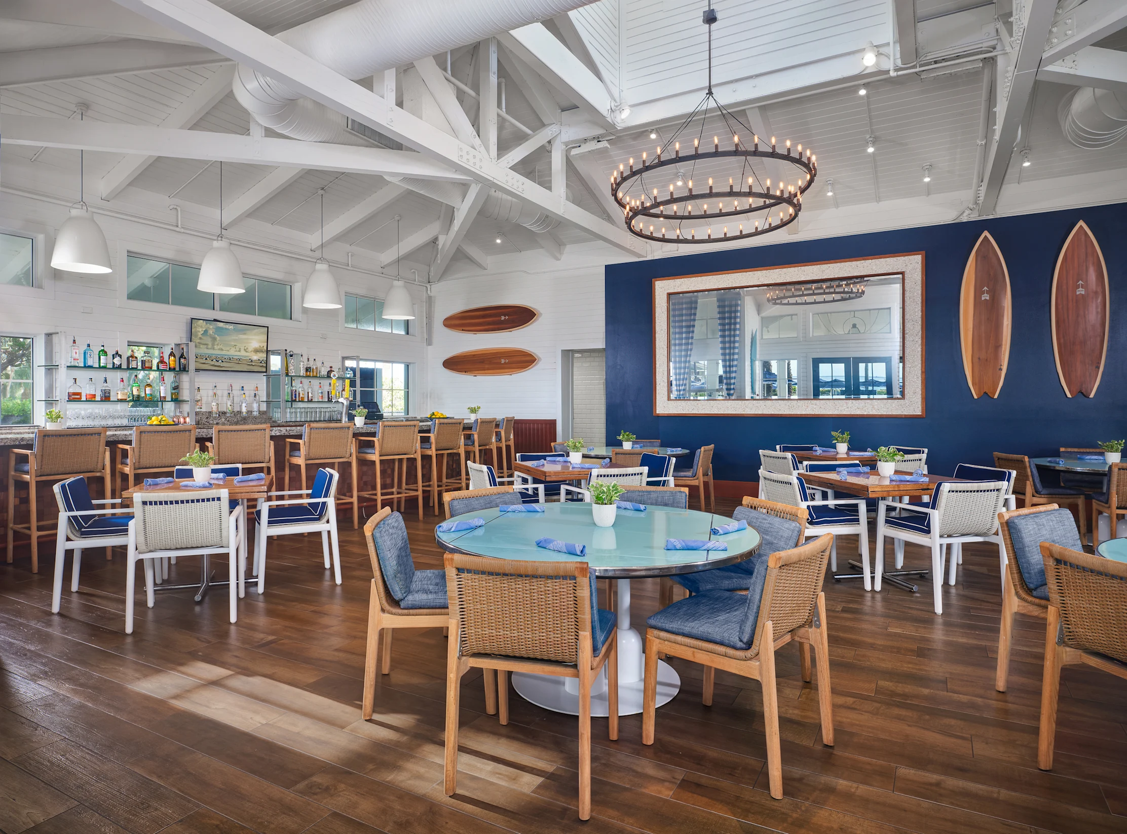 Modern coastal restaurant interior with vaulted ceilings, circular chandelier, bar, coral booth seating, and surfboard decor.