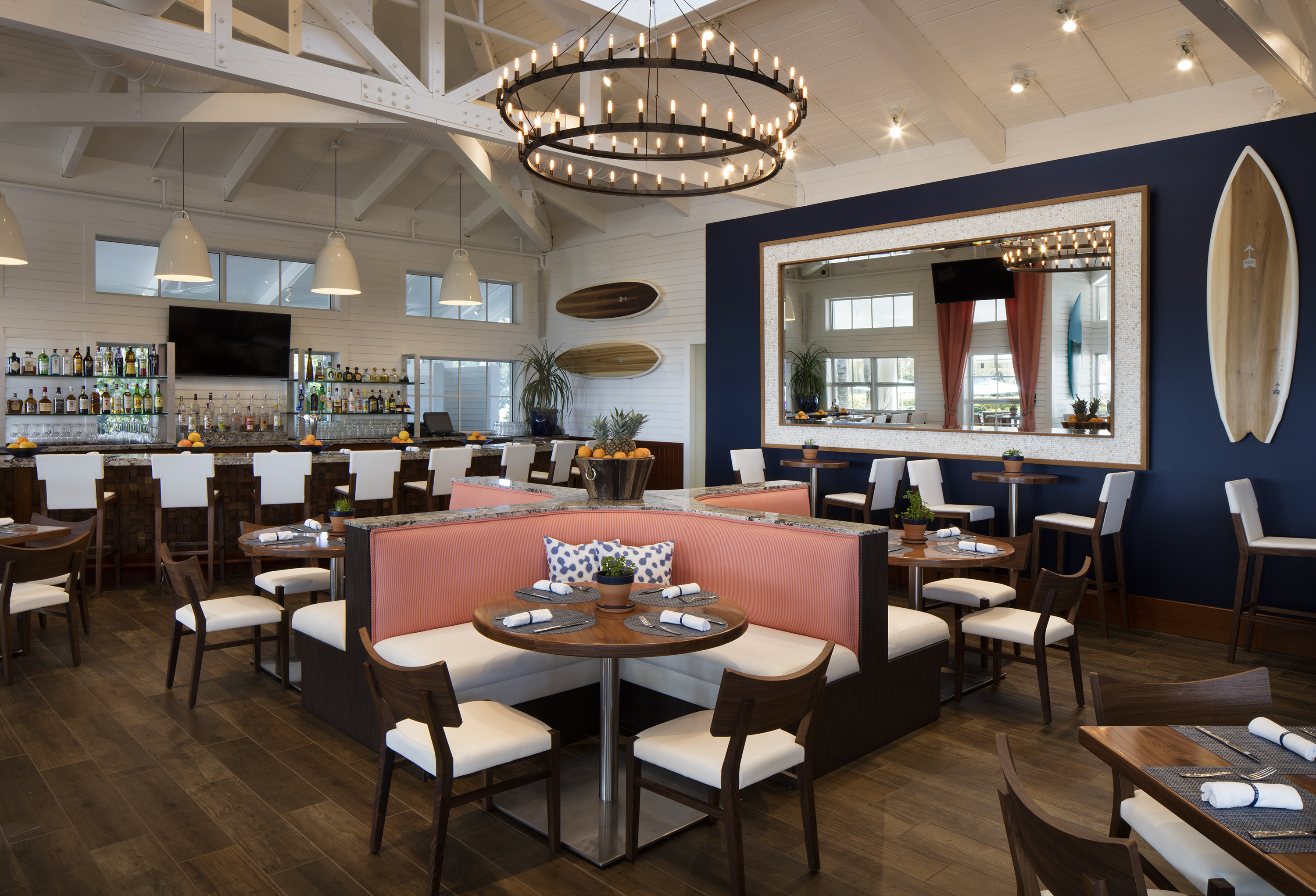 Modern coastal restaurant interior with vaulted ceilings, circular chandelier, bar, coral booth seating, and surfboard decor.