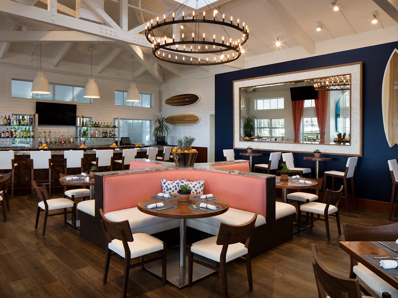 Modern coastal restaurant interior with vaulted ceilings, circular chandelier, bar, coral booth seating, and surfboard decor.