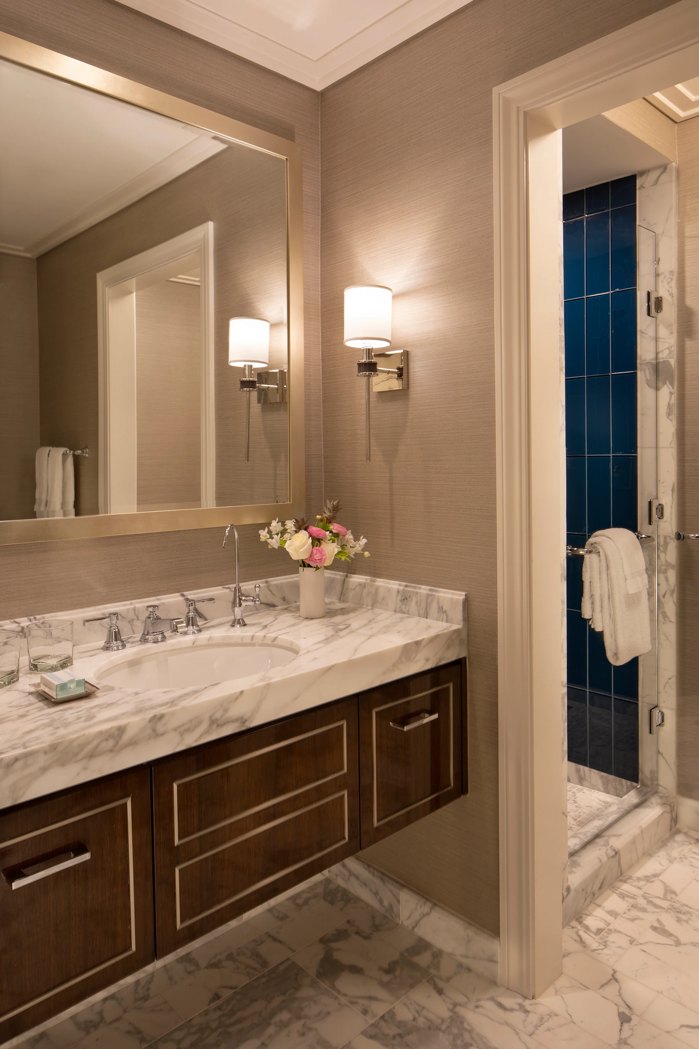 Elegant bathroom with marble vanity, wall sconces, large mirror, and blue tiled shower visible.
