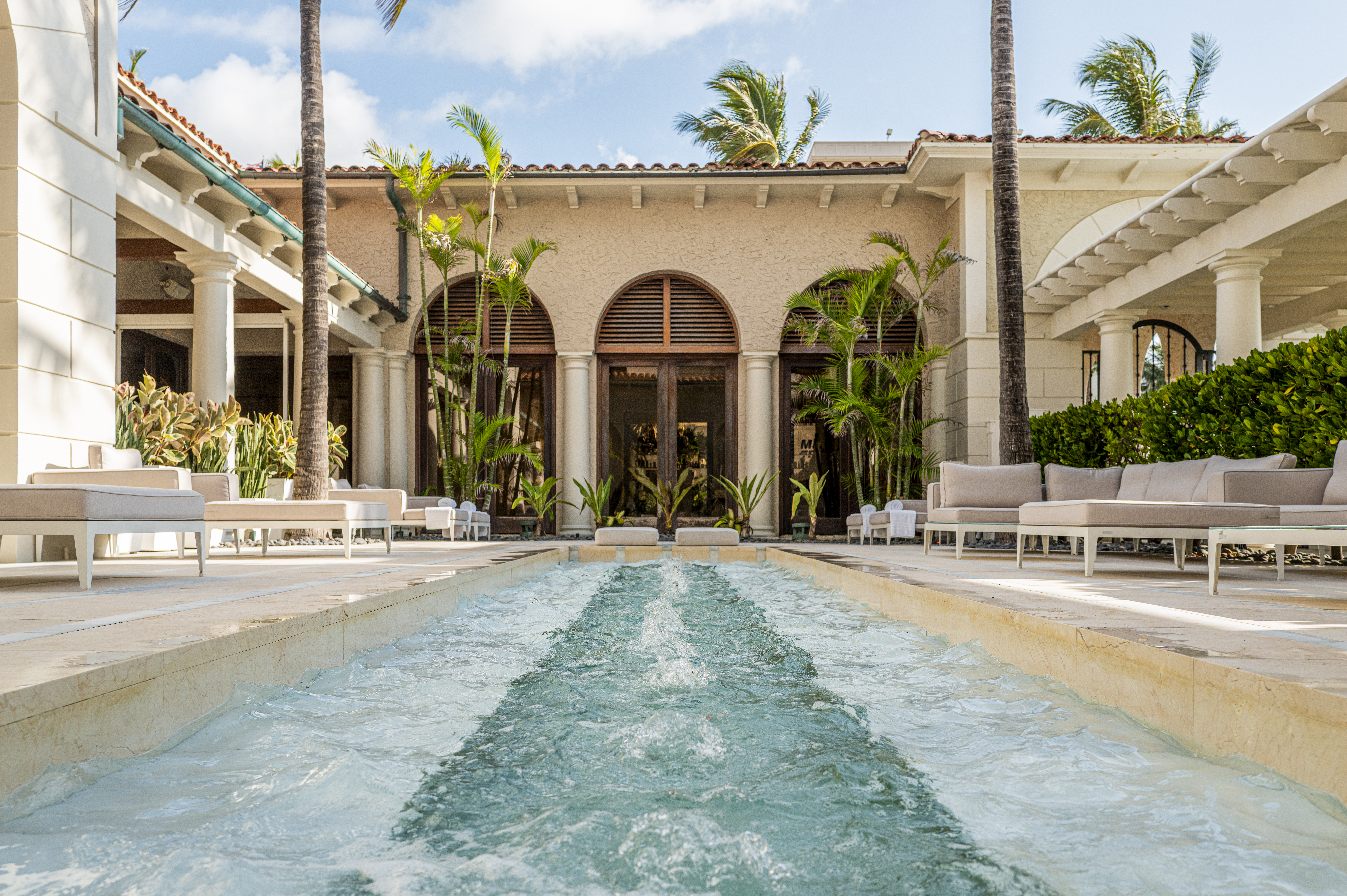 Luxury Mediterranean-style courtyard with turquoise pool, white lounge furniture, palm trees, and arched doors.