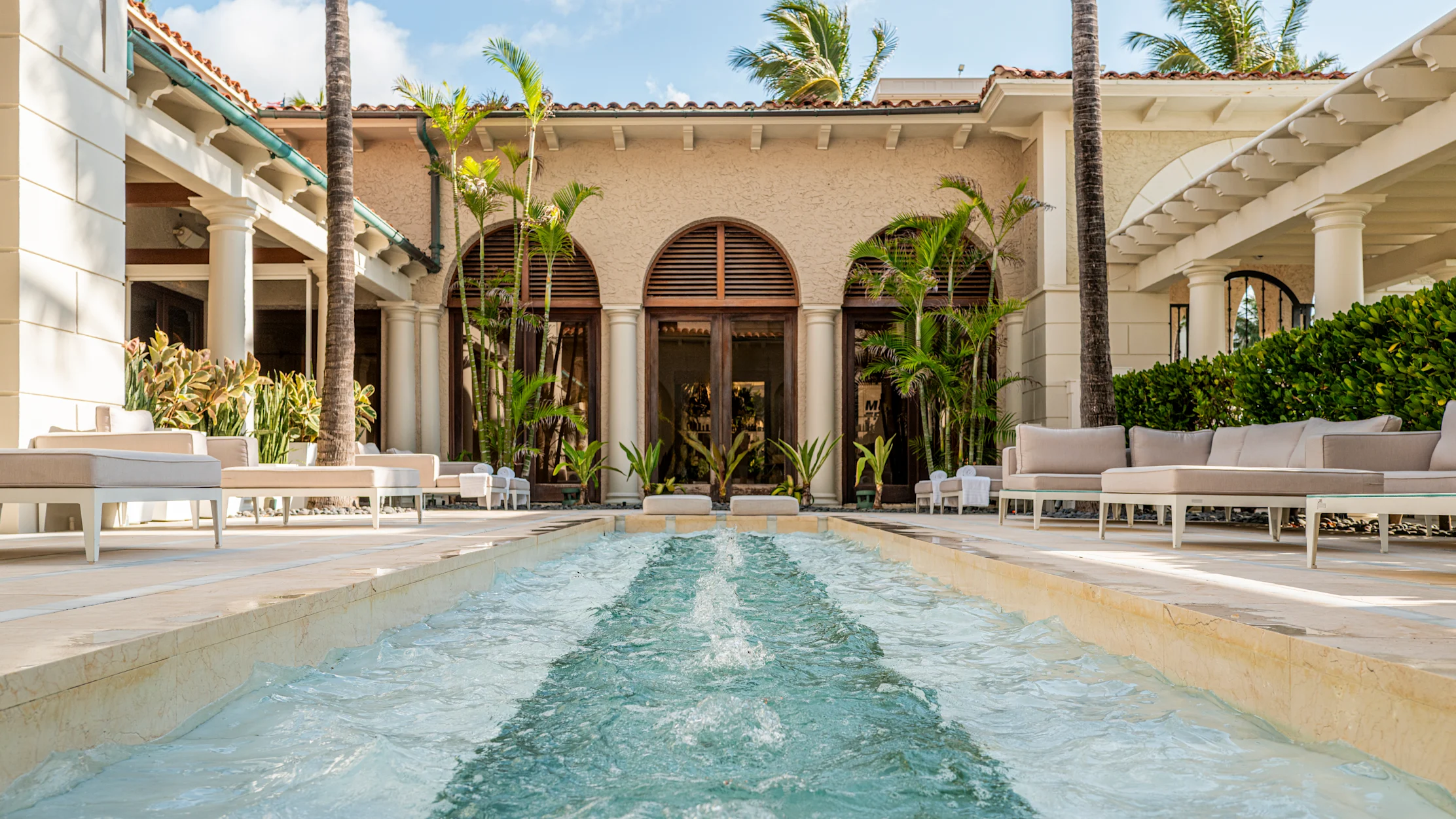 Luxury Mediterranean-style courtyard with turquoise pool, white lounge furniture, palm trees, and arched doors.