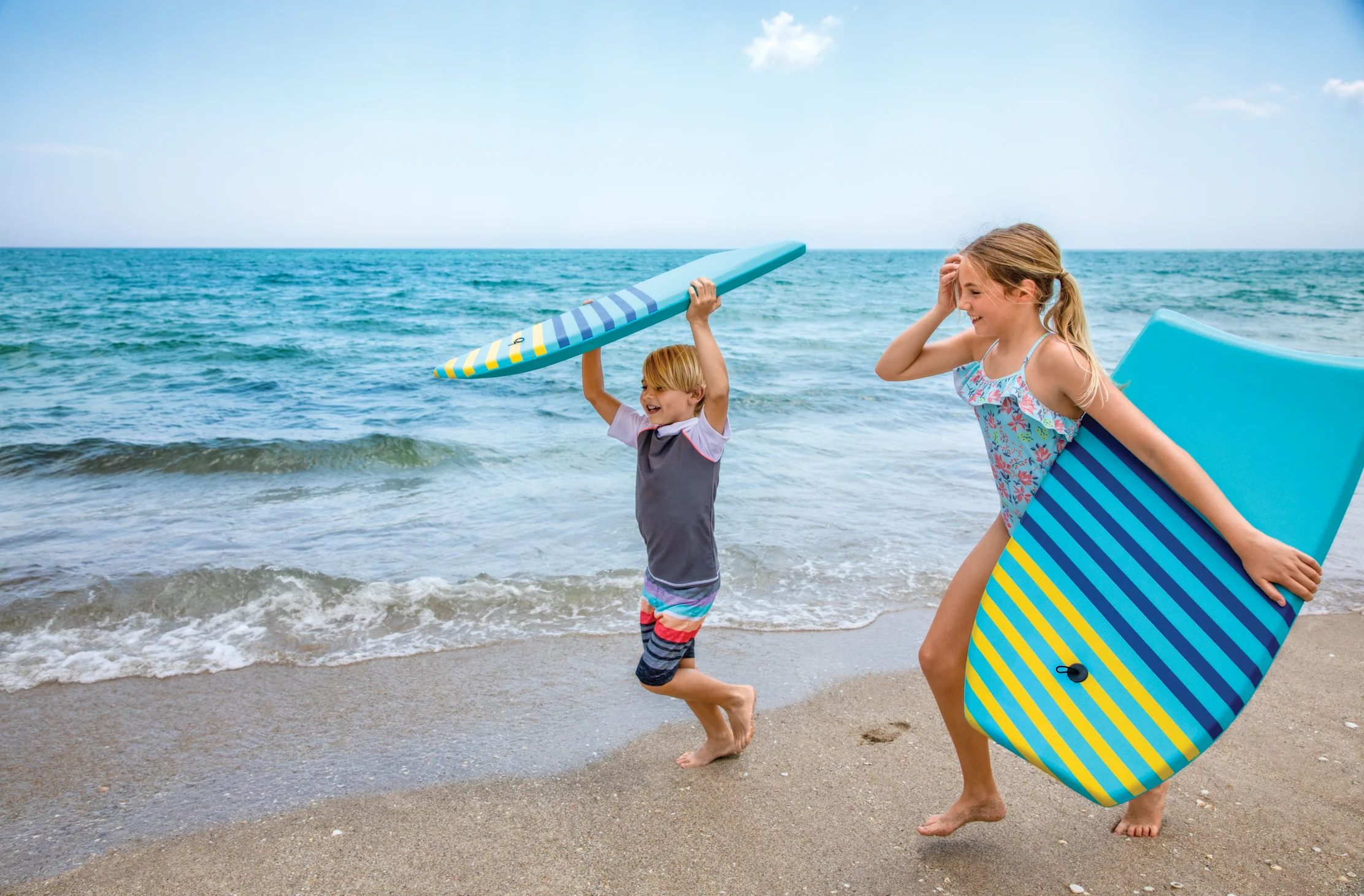 Two children carrying colorful striped boogie boards on a sunny beach with turquoise ocean water.