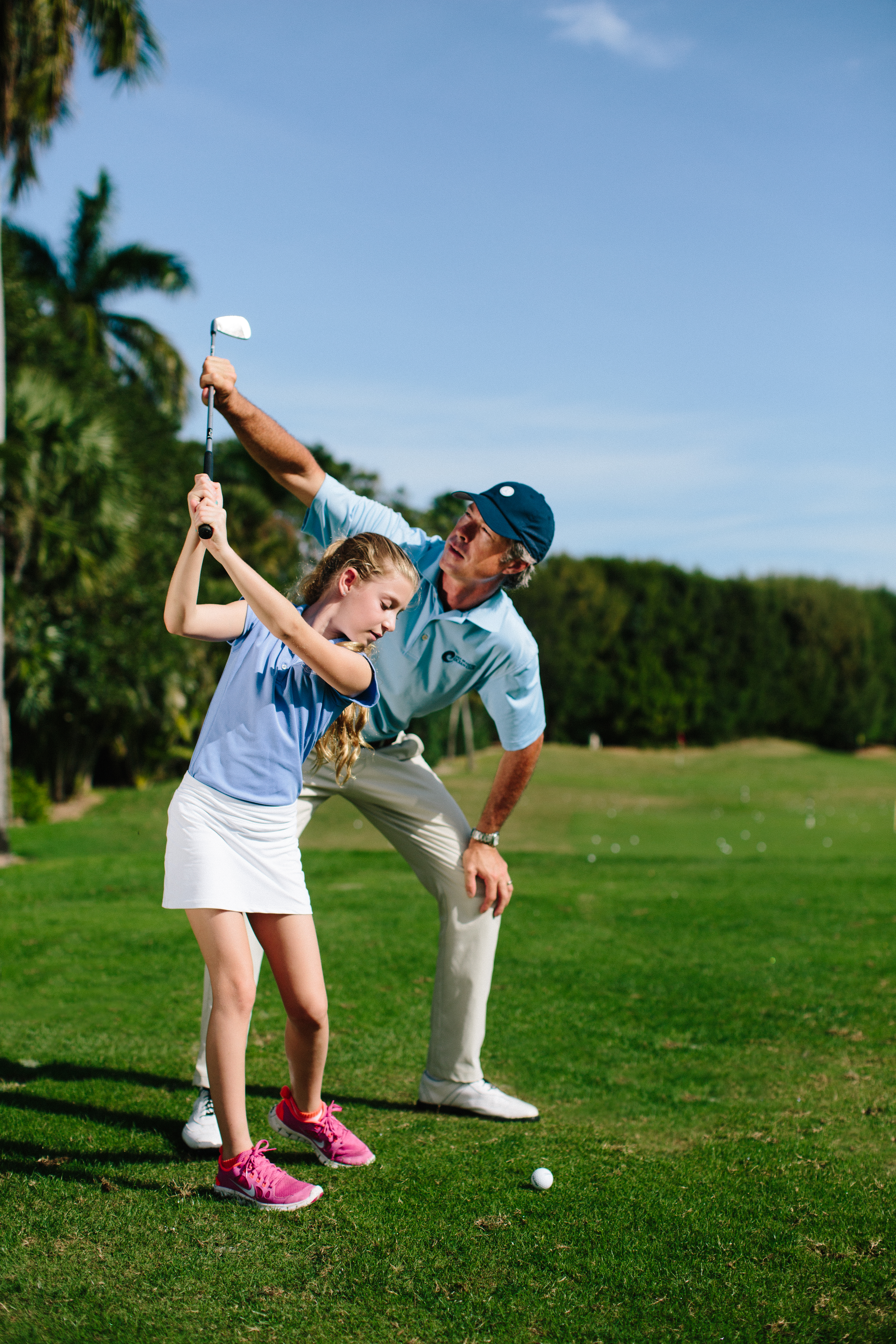 Golf instructor teaching young girl proper swing technique on tropical course with palm trees.