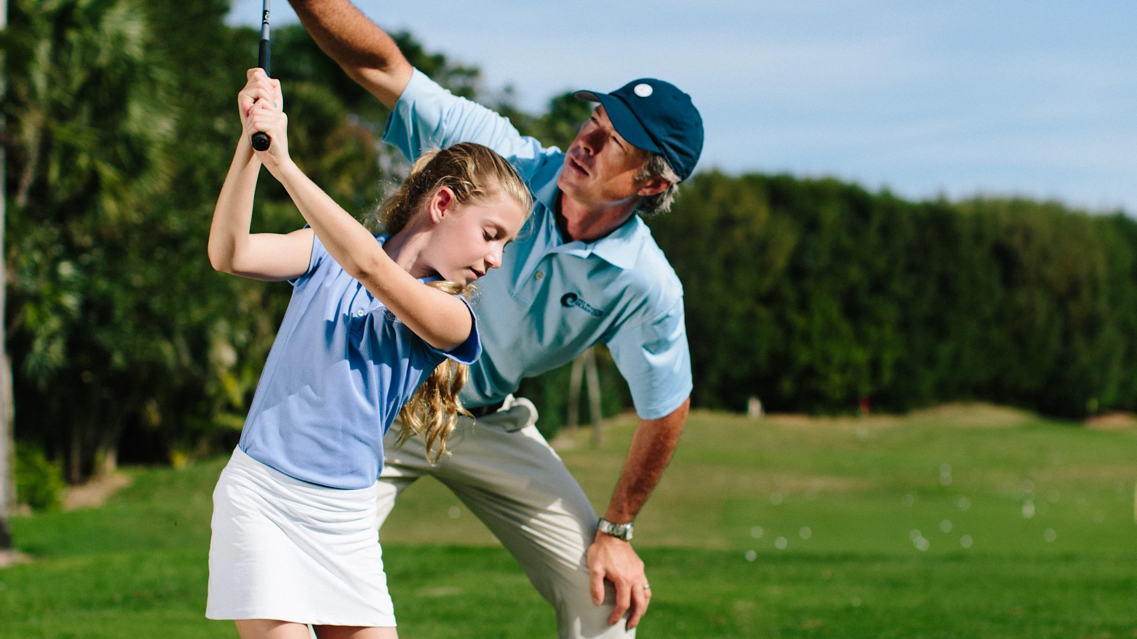 Golf instructor teaching young girl proper swing technique on tropical course with palm trees.