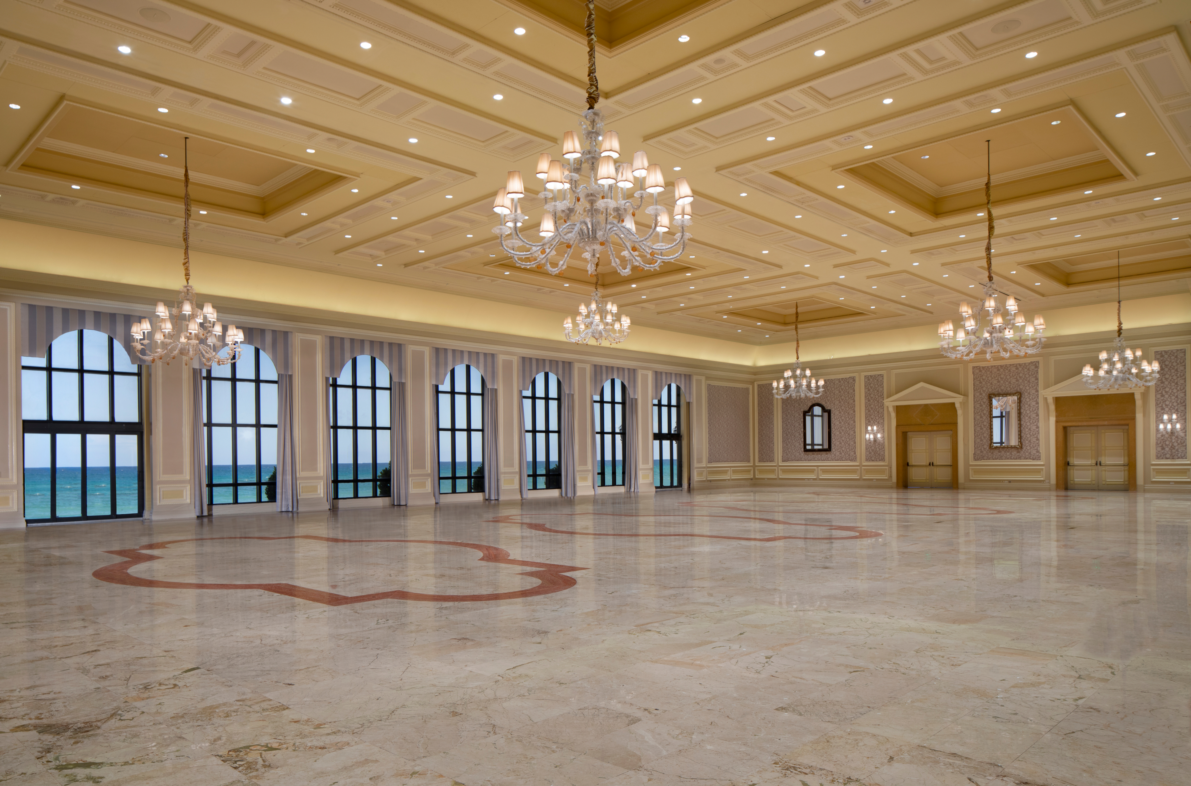 Elegant ballroom with ornate coffered ceiling, crystal chandeliers, marble floors, and arched windows.