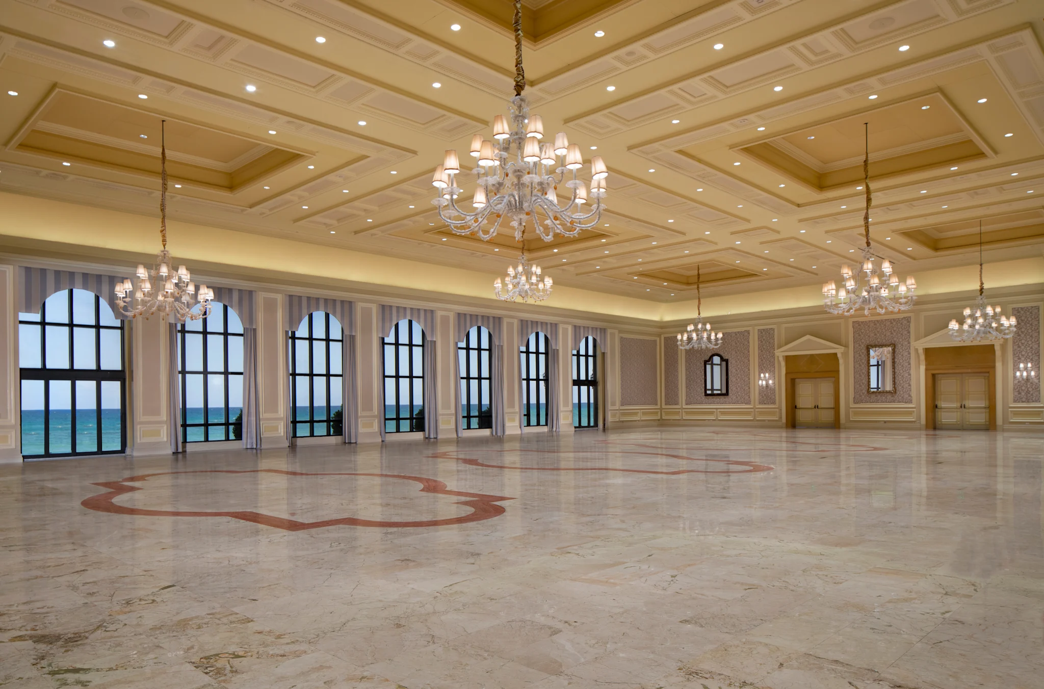 Elegant ballroom with ornate coffered ceiling, crystal chandeliers, marble floors, and arched windows.