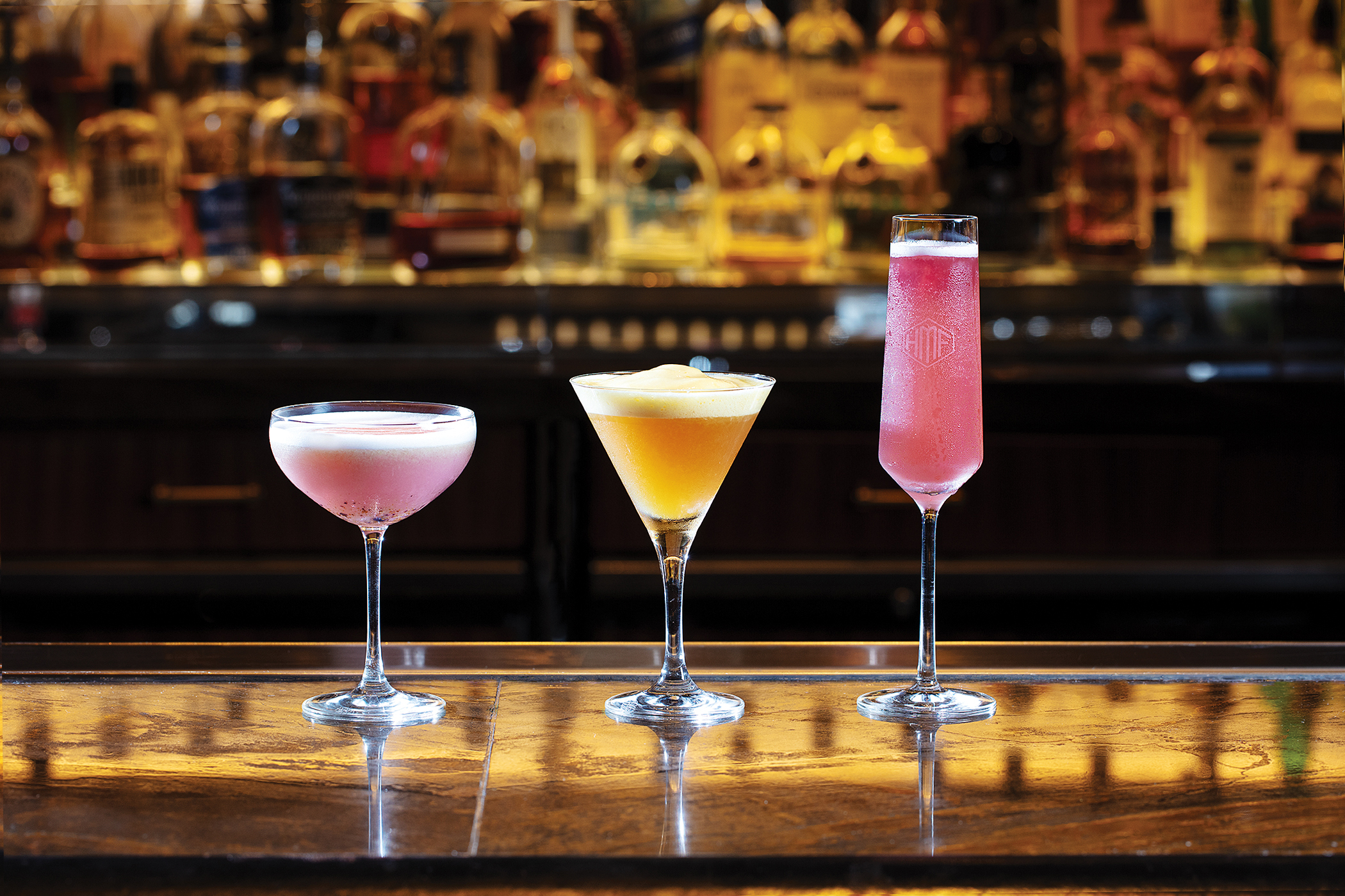 Three colorful cocktails in elegant glasses on a bar with blurred liquor bottles in the background.