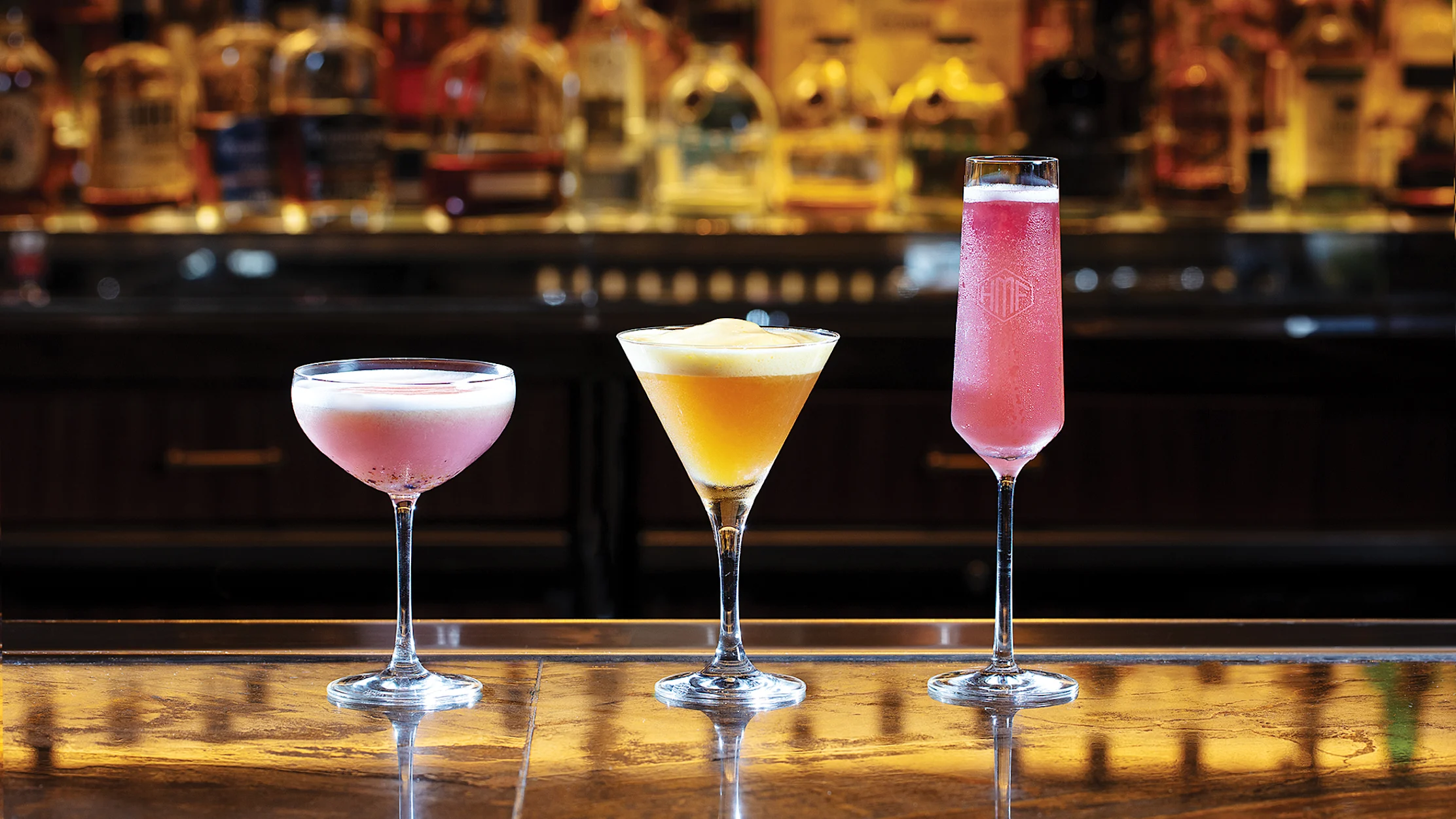 Three colorful cocktails in elegant glasses on a bar with blurred liquor bottles in the background.