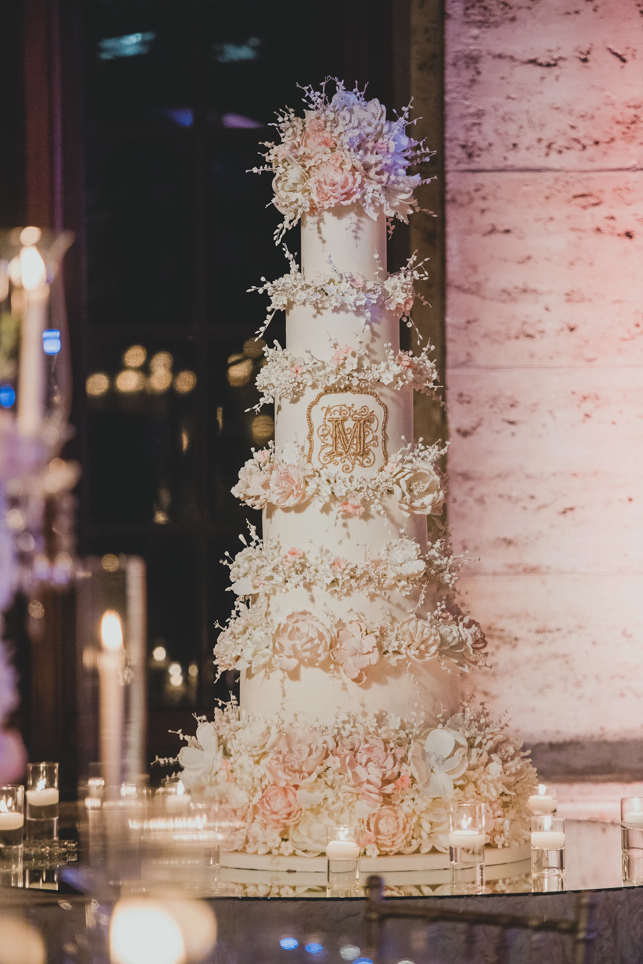 Tall white wedding cake with gold monogram, adorned with cream and blush sugar flowers and candles