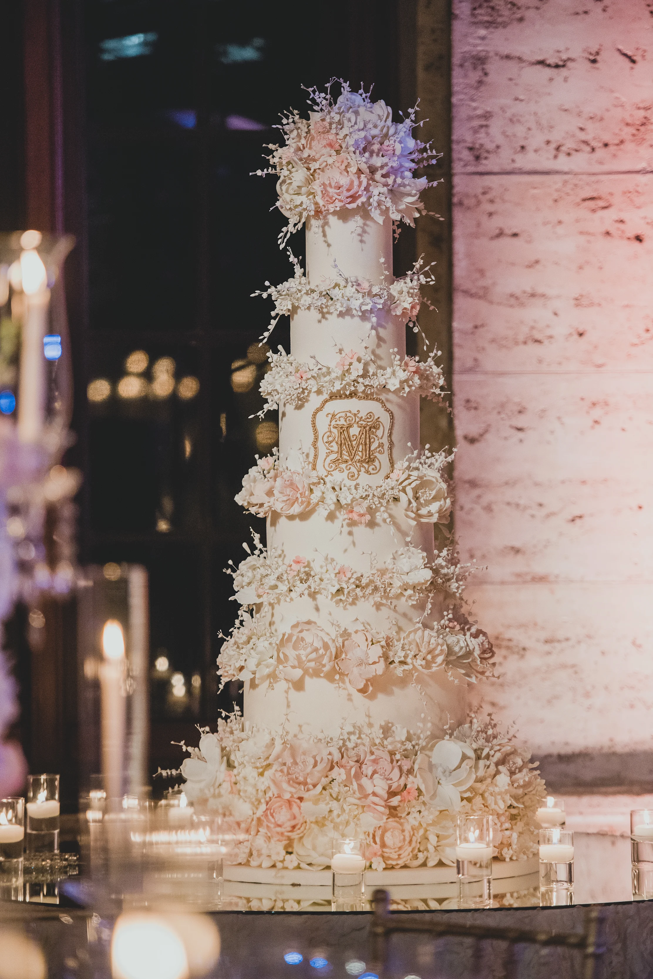 Tall white wedding cake with gold monogram, adorned with cream and blush sugar flowers and candles