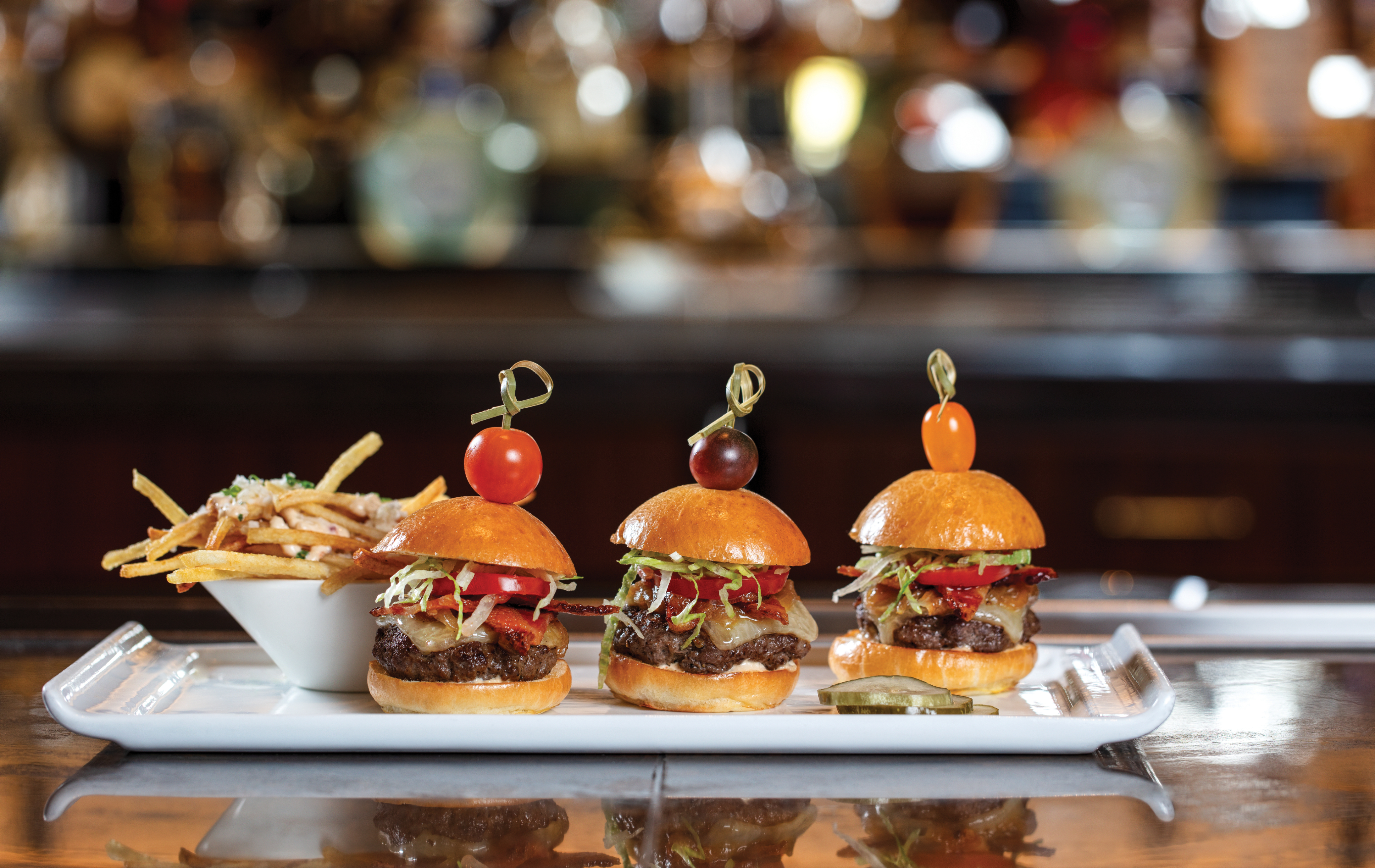 Three mini burgers with tomato garnishes on top, served with fries on a white rectangular plate.