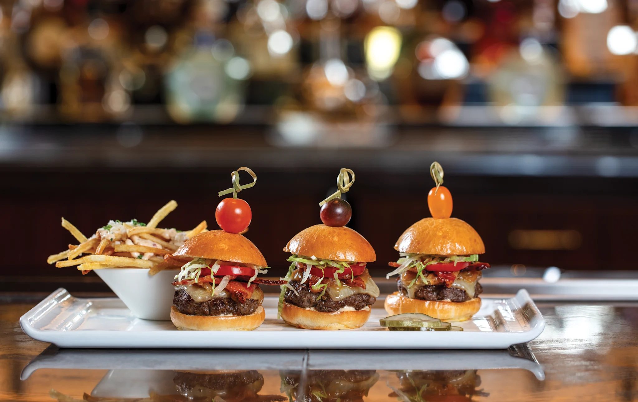 Three mini burgers with tomato garnishes on toothpicks served on a white platter with fries and pickles.