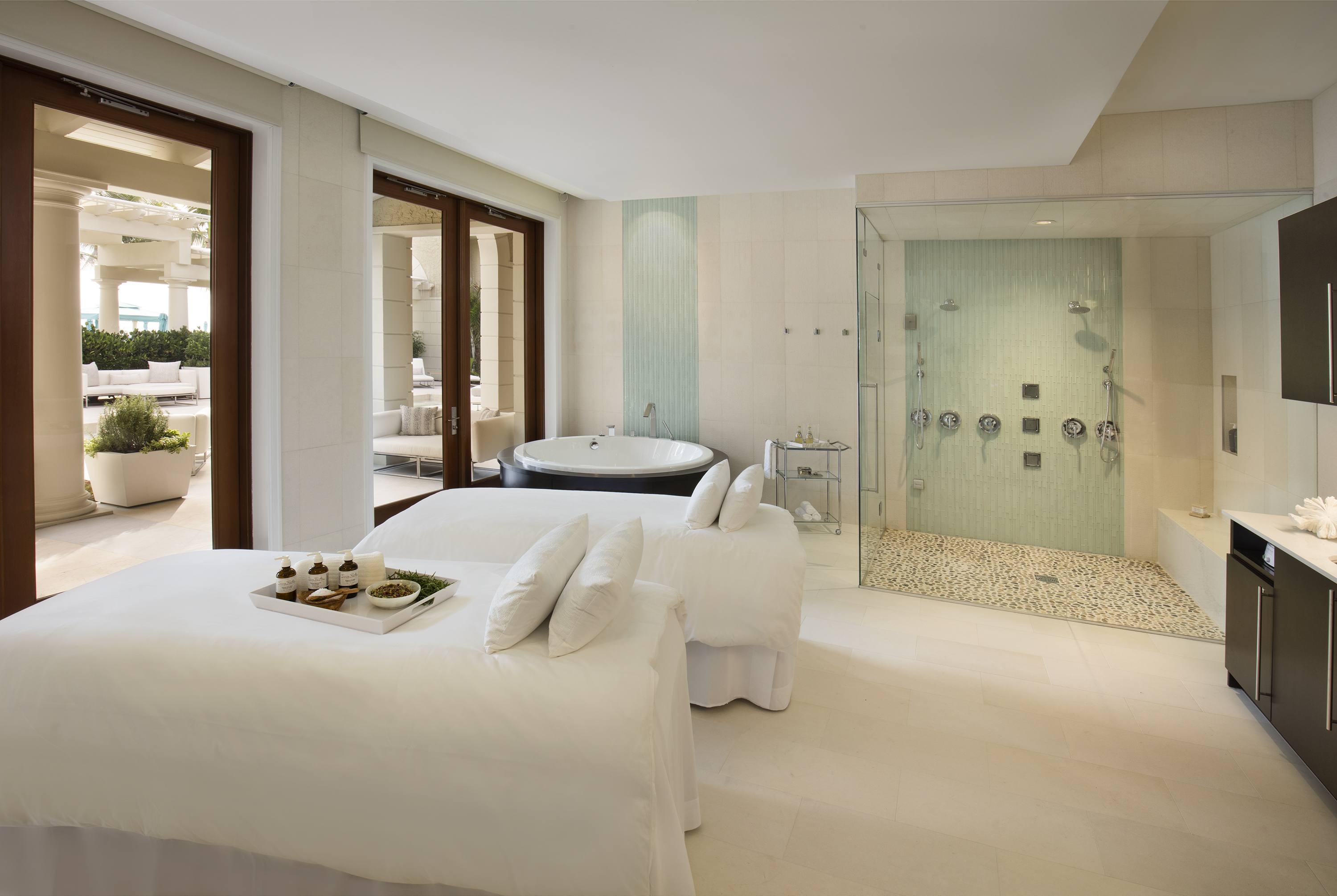 Luxury spa treatment room with white massage bed, glass-enclosed shower, and doors to patio.