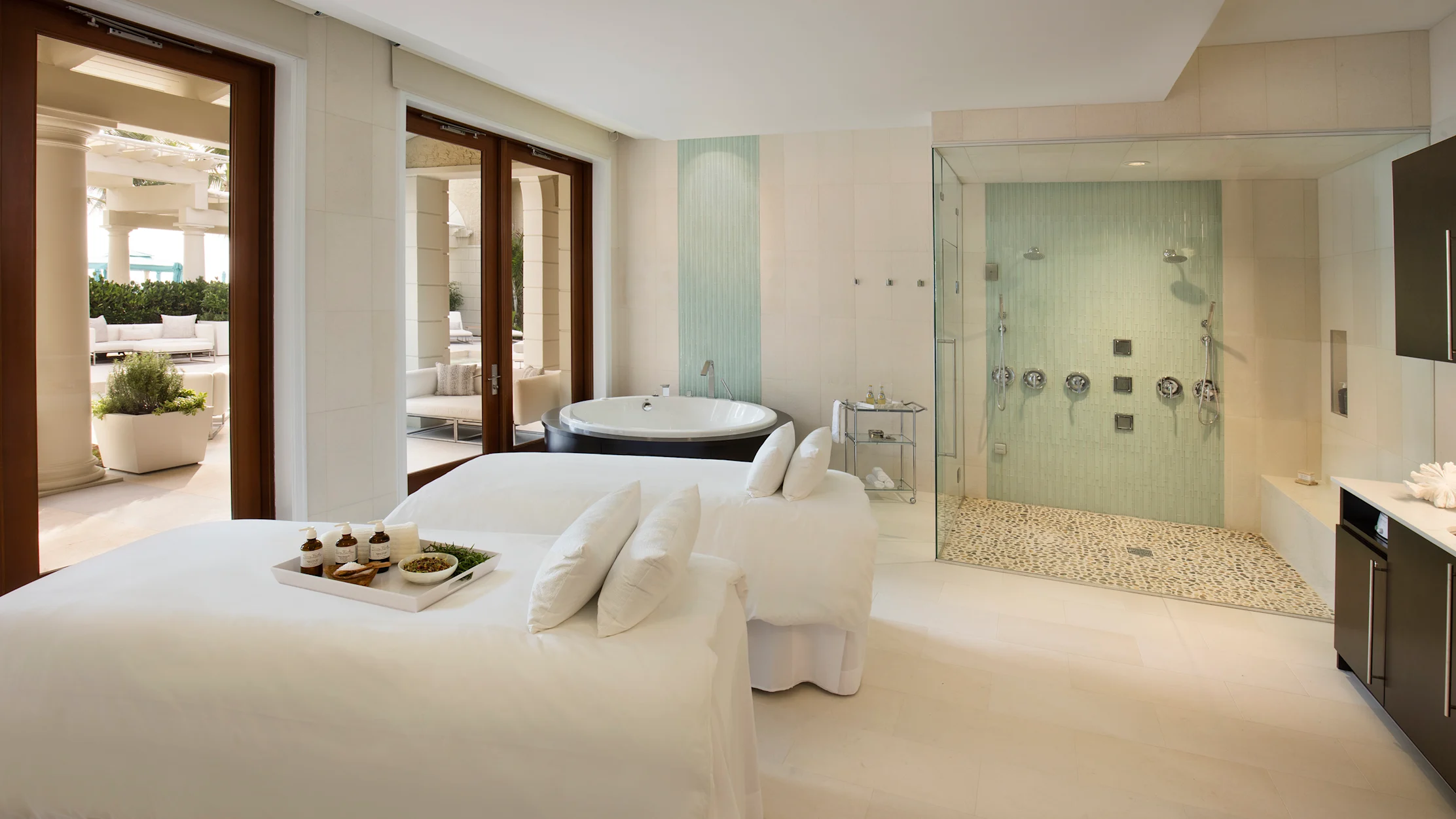 Luxury spa treatment room with white massage bed, glass-enclosed shower, and doors to patio.