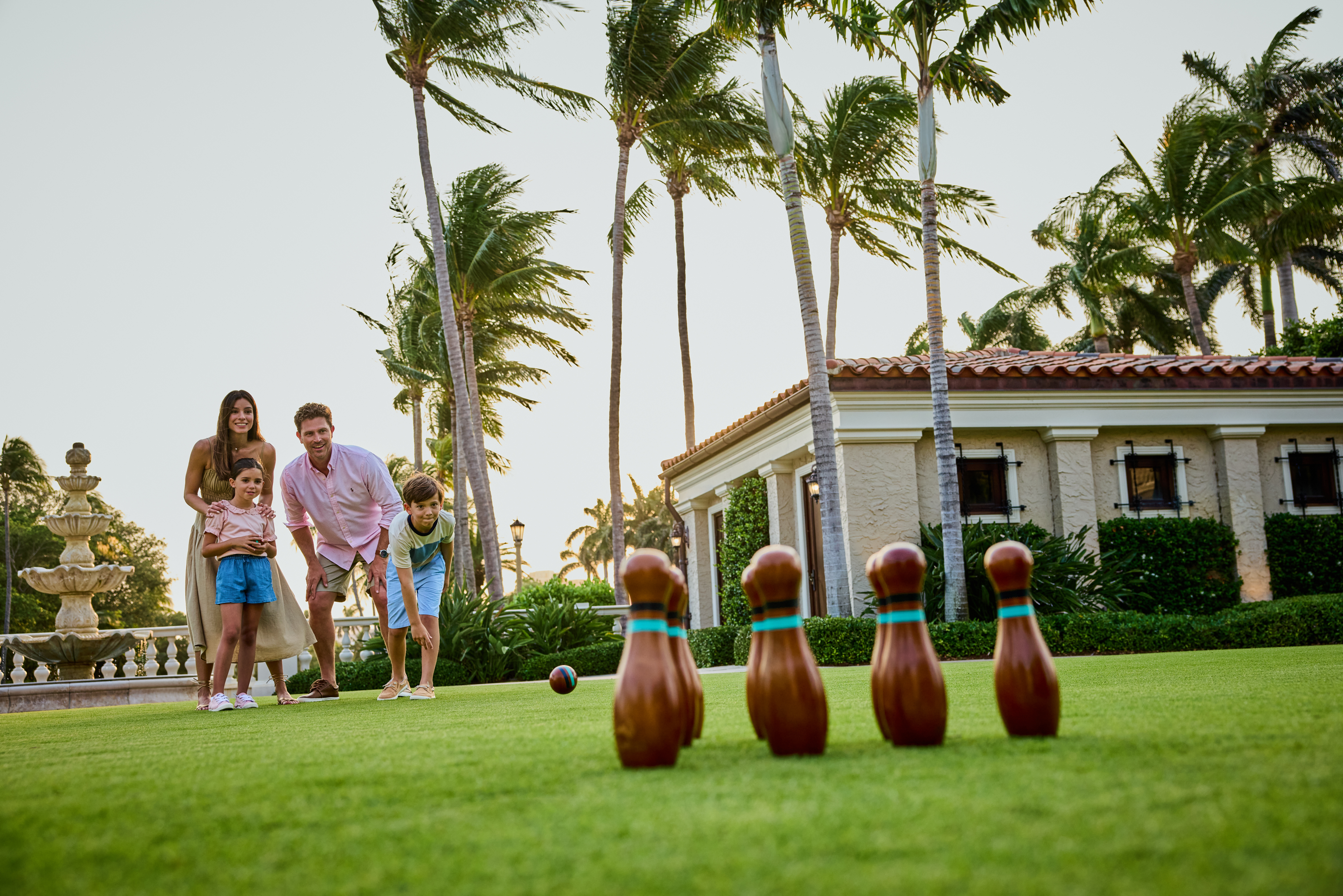 Family of four playing lawn bowling on green grass with palm trees and Mediterranean-style building.