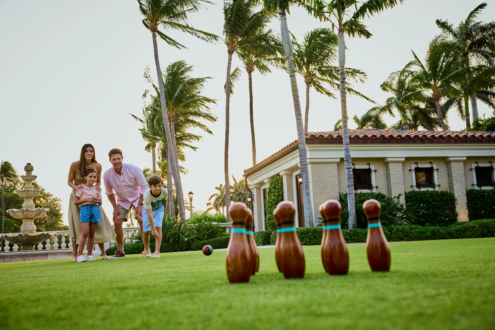 Family of four playing lawn bowling on green grass with palm trees and Mediterranean-style building.