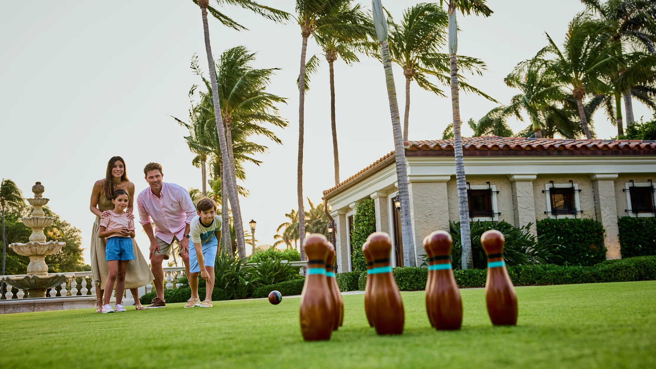 Family of four playing lawn bowling on green grass with palm trees and Mediterranean-style building.